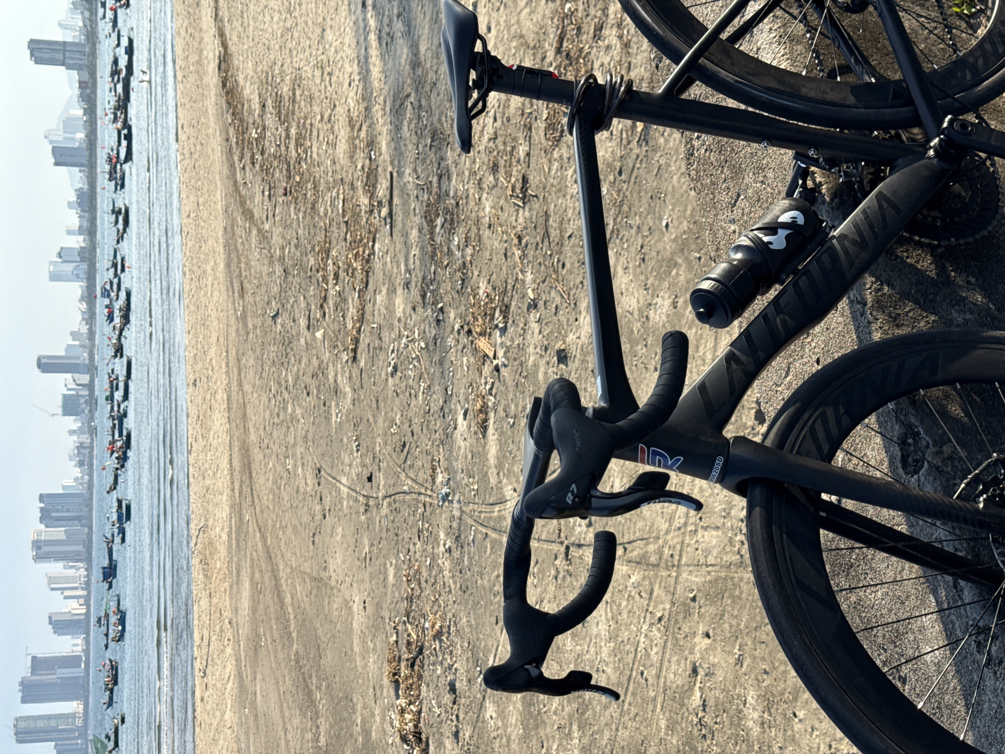 The image shows a black road bicycle lying on its side on a sandy surface, likely a beach. In the background, there is a body of water with several small boats and a city skyline visible in the distance. The scene appears to be in Đà Nẵng, Vietnam, based on the style of the boats and buildings. The bicycle has a water bottle mounted on the frame. The sandy surface is littered with small pieces of debris. The lighting suggests the photo was taken during the day.