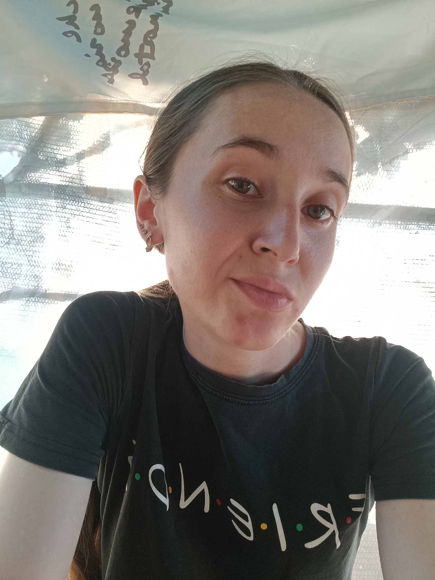 A young woman with brown hair pulled back is taking a selfie, looking directly at the camera. She is wearing a dark t-shirt with the word "FRIENDS" printed on it in white letters, with colored dots between each letter. She has earrings in her left ear, and there appears to be writing on a light-colored fabric in the background, which is illuminated by sunlight. The setting is outdoors, likely in the city of Vypasne, Ukraine.
