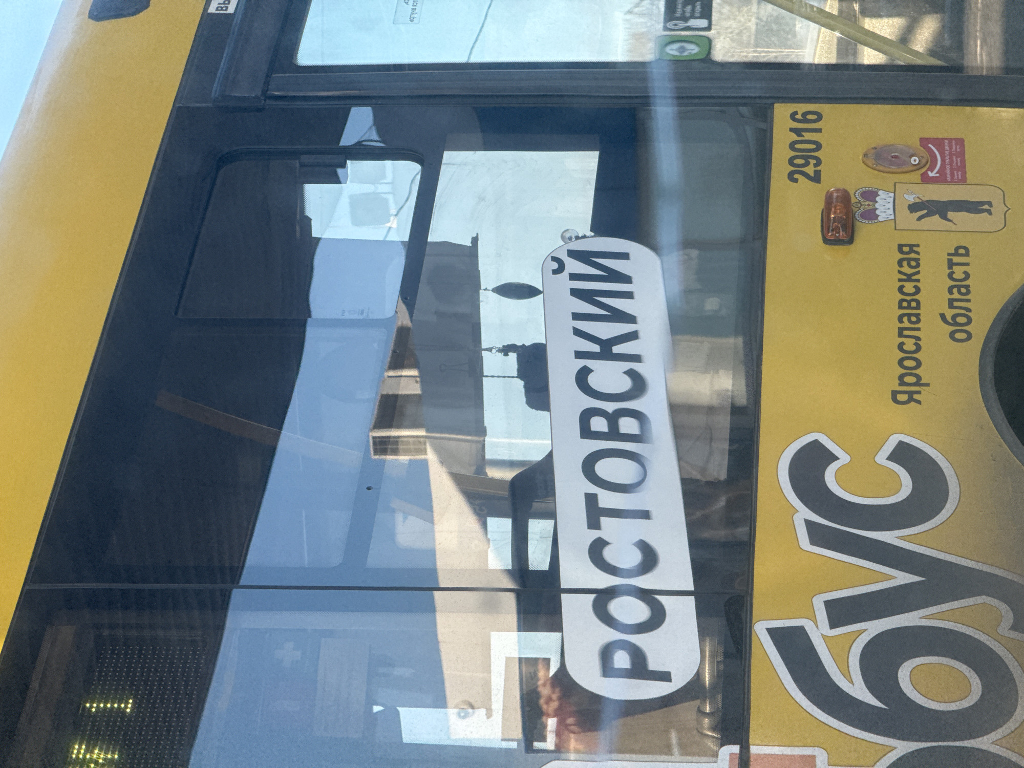 The image shows a section of a yellow bus in Ново-Никольское, Russia. Visible on the side of the bus is the word "Ростовский" (Rostovsky), which is a common surname and place name. Also on the bus is information stating that it is from the Yaroslavl region, identified by its coat of arms and the text "Ярославская область" (Yaroslavskaya oblast'). The number "29016" is printed above the region's coat of arms. The image includes a window of the bus that reflects the surroundings.