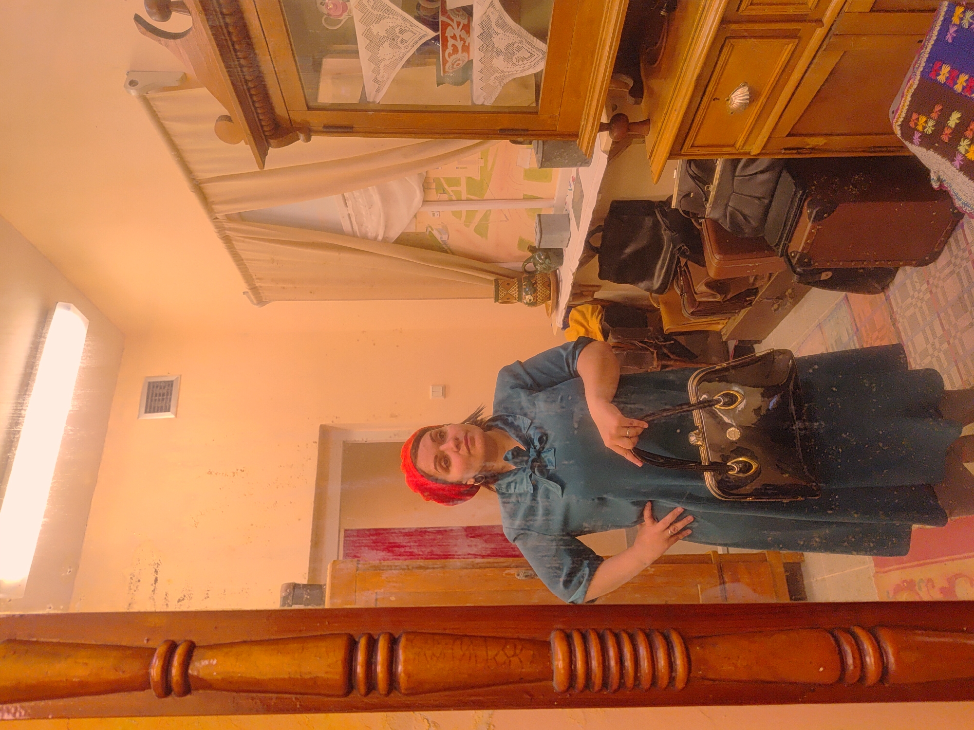 The image depicts an indoor scene with a woman centrally positioned, holding a black patent leather handbag. She wears a red beret and a teal-colored dress or tunic with a bow at the neck. She has dark hair and a neutral expression, looking directly at the viewer. Rings are visible on her fingers.
The setting appears to be a room or hallway with a distinct vintage aesthetic, consistent with a traditional home or museum in Russia. The walls are a warm, light orange color, showing some signs of age or wear. In the background, there's a wooden display cabinet or hutch with glass doors, containing white lace doilies and other decorative items. Below the cabinet, a collection of assorted suitcases and bags, varying in material and size, are stacked, suggesting storage or readiness for travel.
To the far right, a colorful embroidered textile, possibly a rug or blanket, adds to the traditional decor. On a table beneath the cabinet, ceramic teacups and pots are visible. A map or faded painting is on the wall behind this arrangement. A long fluorescent light fixture illuminates the scene from the left, and a small square vent is on the wall. In the foreground, a detailed wooden balustrade or railing is prominent, implying a staircase or elevated section. A doorway is visible in the background, to the right of the woman. No specific activity is taking place other than the woman posing. There is no visible text. The overall atmosphere is reminiscent of a bygone era.