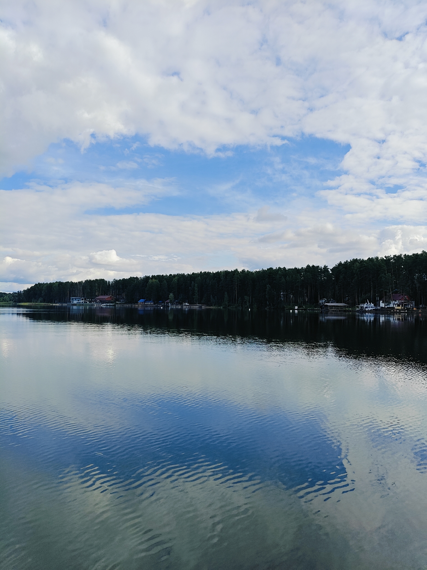 The image shows a wide, calm body of water, likely a river or lake, with a dark line of trees forming the horizon. Several buildings, including what appear to be houses and boathouses, are visible nestled within the treeline. A few boats are docked along the shore. The sky is partly cloudy, with patches of blue visible between the white clouds. The water reflects the sky and the trees, creating a mirror-like effect with subtle ripples distorting the reflections. The scene suggests a tranquil, natural environment possibly a resort or residential area along the water. The location is the Kama River in Chaykovsky, Russia. There are no people visible in the image.