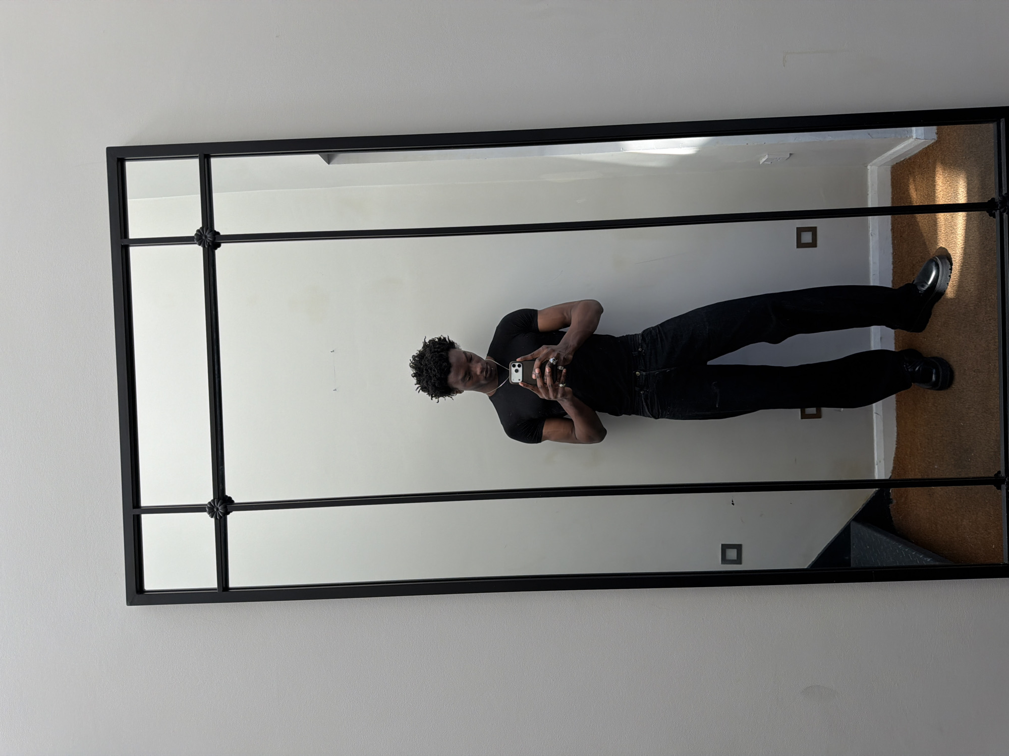 A young Black man is captured taking a mirror selfie in an indoor setting. He stands upright, centered within a large, rectangular mirror featuring a sturdy black metal frame. The frame is designed with horizontal and vertical crossbars, creating multiple pane-like sections, and a decorative rosette ornament is visible at the central intersection of these bars.
The man is dressed entirely in black, wearing a short-sleeved t-shirt, trousers, and polished black shoes with thick soles. His hair is short and dark with a curly texture. He accessorizes with a necklace and several rings on his fingers, particularly on his right hand. He holds a white smartphone with multiple camera lenses at chest level, aiming it at his reflection to take the photo.
The background visible in the mirror's reflection consists of plain, light-colored walls, featuring two small, dark square elements that could be light fixtures or outlets. The reflected floor is covered with a warm, light brown or beige carpet, upon which a distinct rectangular patch of sunlight is visible, suggesting daylight entering from an unseen window. A dark, angled structure at the lower right of the reflection hints at a staircase or step. The mirror is mounted on a similar light-colored wall. The scene conveys a casual, everyday moment, with the man focused on his phone. There is no visible text in the image.