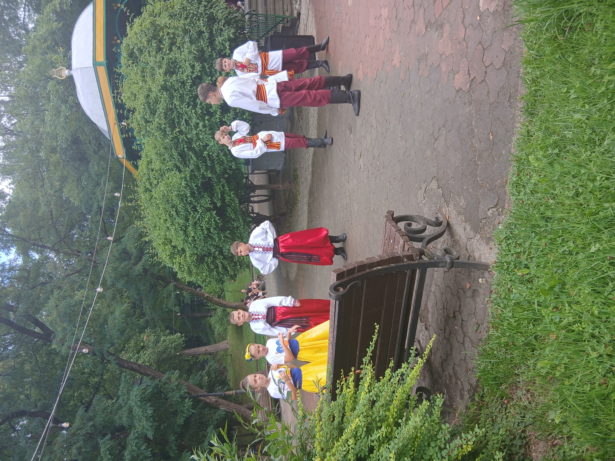 A group of six young people in traditional Ukrainian folk costumes stand in a park. The two girls on the left wear white tops with blue and yellow accents and yellow skirts, while the four boys wear white shirts with red embroidery, red sashes, and red pants tucked into black boots. They are standing on a paved walkway in a park with lush green trees and bushes surrounding them. In the background, a white and green gazebo with a lyre-like ornament on top is visible. The ground is paved with interlocking stones. The overall scene suggests an outdoor performance or cultural event in Truskavets, Ukraine.