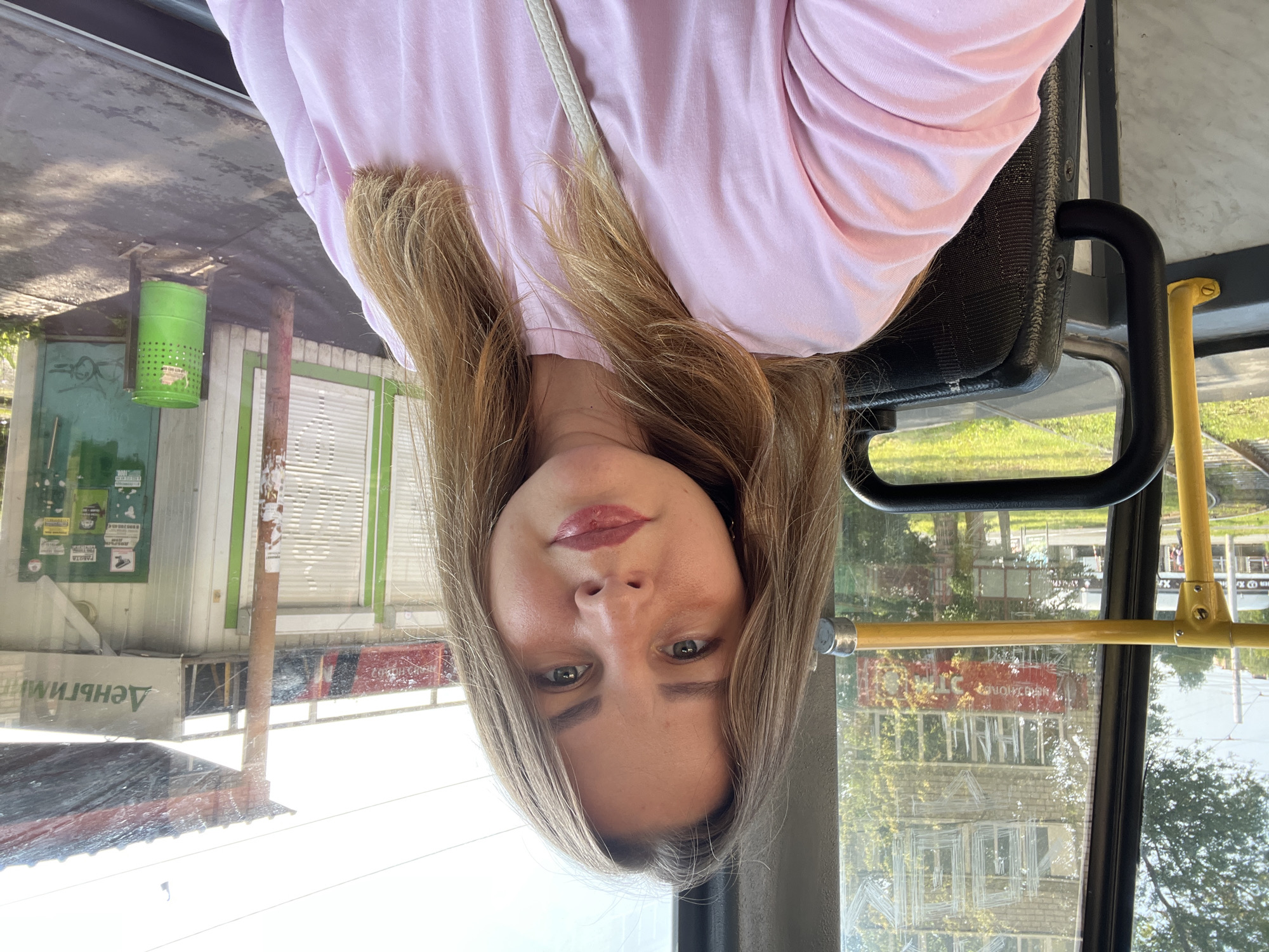 The image is a selfie taken inside a public transportation vehicle, likely a tram, in Izhevsk, Russia. The woman in the foreground is wearing a pink t-shirt and has blonde hair. She is looking at the camera with a neutral expression.
Through the window, we see a typical urban landscape. A building with several floors is visible, featuring windows and some signage. The building appears to be older, with a facade that suggests a Soviet-era construction. Outside, there's greenery, indicating it's daytime and likely summer. Another structure with a sloped roof and various posters or advertisements is also present. A bright green trash can stands near this structure. The presence of tram tracks suggests the vehicle is on a route. The overall impression is a snapshot of everyday life in a Russian city.