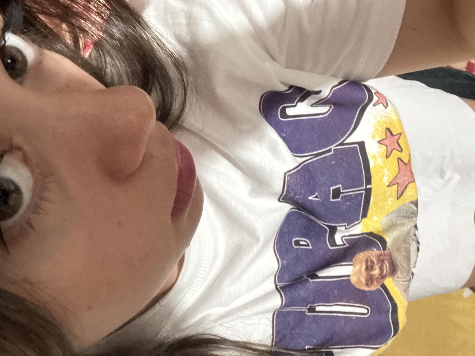 The image is a close-up, overhead shot of a young woman wearing a white t-shirt with a graphic print. The t-shirt features large purple lettering, partially visible, and a smaller image of a man's face, possibly Tupac Shakur, on a yellow background with red stars. The woman's face is visible from the nose up, with her brown eyes looking slightly off-camera and her lips closed. Her dark brown hair is partially visible. The background is blurred and appears to be a solid yellow surface. There is no discernible location in the city of Garmen, Bulgaria.