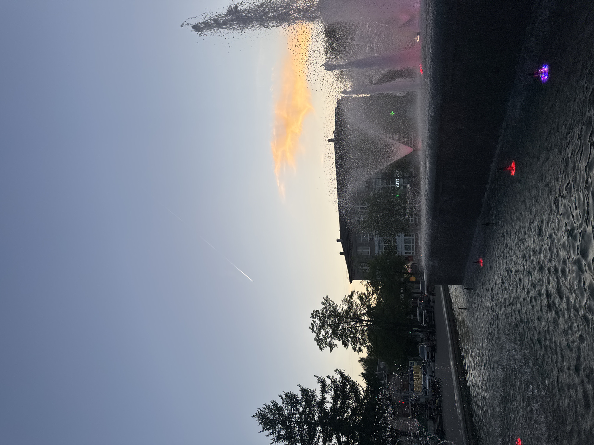 The image shows a view of a water fountain in Chaikovsky, Russia at dusk. The sky is light blue with an orange cloud near the center. A building is visible in the background behind trees. There are red, purple, and green lights illuminating the fountain. Water is flowing over the edge of the fountain, creating a textured, reflective surface. A contrail of a plane cuts diagonally across the sky.