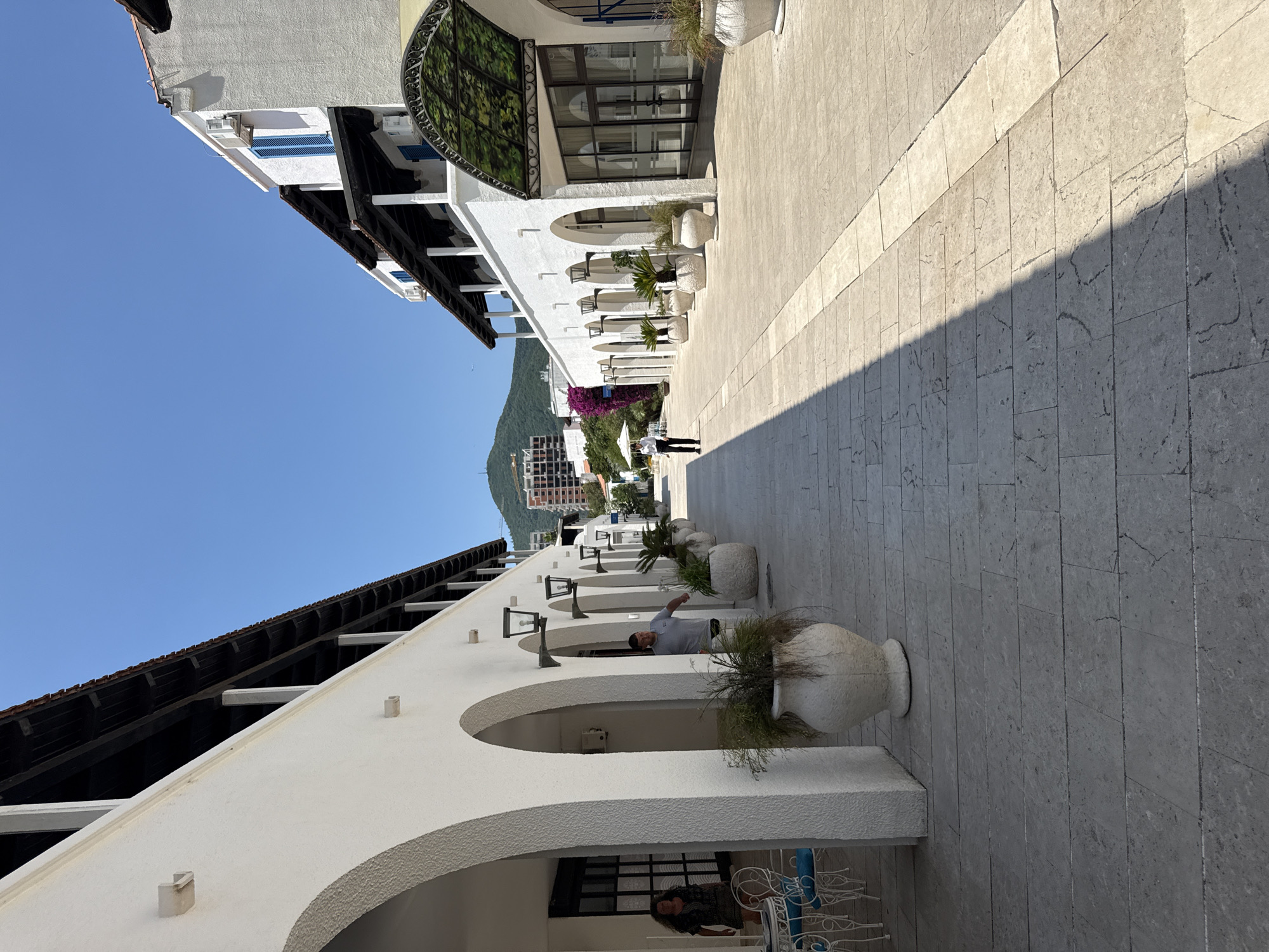 This is a vertical, eye-level outdoor shot of a walkway with a building on the left and a paved area on the right. The sun is casting a distinct shadow across the paved walkway, suggesting it is daytime. The sky is a clear, vibrant blue, indicating good weather.
The building on the left features white stucco walls and arched openings, with dark wooden beams supporting a sloping roof. Several large, white planters with greenery are placed along the walkway. A man in a light gray shirt and dark pants is visible standing near one of the planters, pointing towards the distance. Further down the walkway, a person with dark hair is seated at a table with a white wrought-iron chair. The architectural style suggests a resort or hotel setting, possibly in a Mediterranean or coastal region. In the background, a lush green mountain slopes down towards buildings, and there are some purple flowers adding a touch of color.
The paved walkway is made of light-colored rectangular stones. The overall scene conveys a sense of leisure and a pleasant, sunny day. No visible text is present in the image.