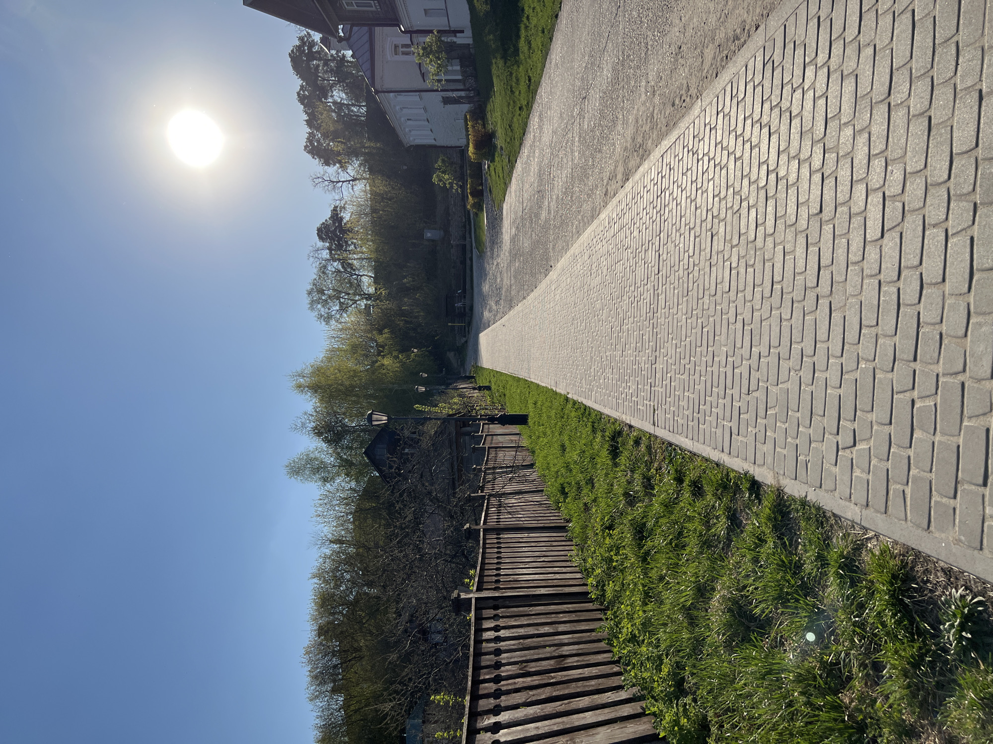 The image depicts a pathway in Sviyazhsk, Russia. The pathway is constructed of gray pavers, bordered by a strip of green grass. To the left of the grass is a wooden fence. Beyond the fence are trees and the clear blue sky, with the sun shining brightly. On the right side of the pathway are more trees, a patch of green grass, and a building in the background. The image is taken at a low angle and the sun is visible in the sky.