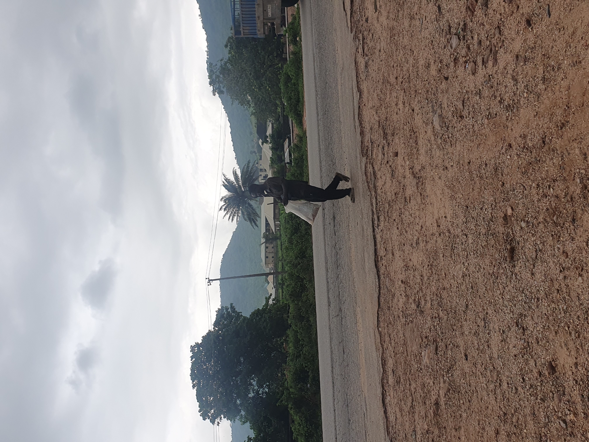 A Black man, wearing a dark shirt and dark pants, walks on the side of a two-lane asphalt road carrying a large, white plastic bag in his right arm. The road is cracked and has a gravel shoulder. In the background, there is a dense green bush and several buildings, including a light blue truck with a blue grate on the side. Further in the distance, hills covered in trees and vegetation are visible under a cloudy sky. The scene appears to be in Lokoja, Nigeria.