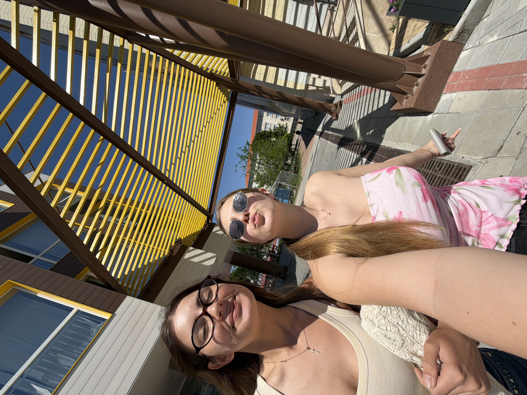 Two young women are taking a selfie outdoors. The woman on the left is wearing glasses and a light-colored tank top, with a necklace. The woman on the right is wearing sunglasses and a strapless pink floral top. They are standing in front of a modern building with yellow and brown architectural elements. The background shows trees, other buildings, and a clear blue sky. The location is identified as Burgas, Bulgaria. The ground is paved with square tiles, and there are metal structures and planters. The lighting suggests it is a sunny day.