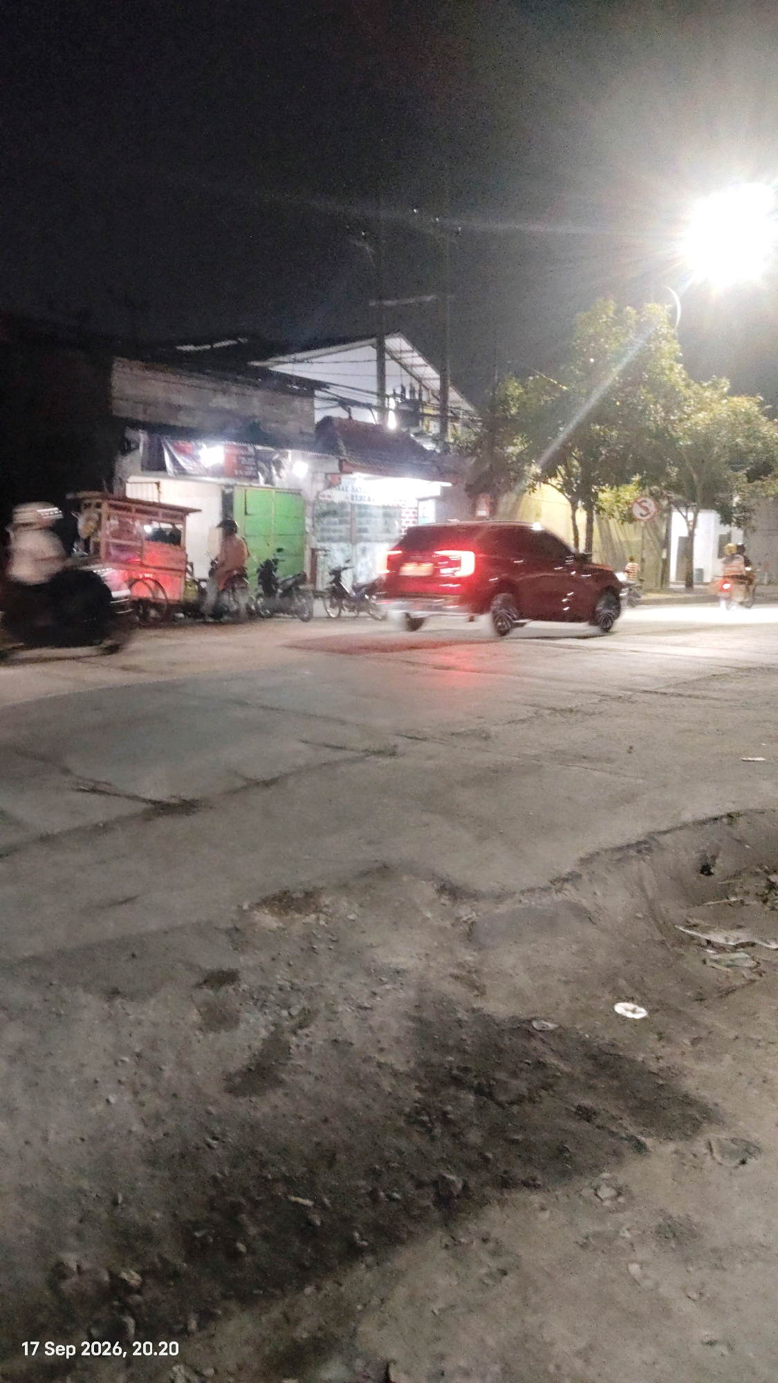 A red SUV is turning left at an intersection at night, with its brake lights illuminated. The SUV is in the middle ground, facing left. To the left of the SUV, there is a food cart, several motorcycles, and a person on a motorcycle. To the right of the SUV, there is a street lamp, trees, and a sidewalk with some buildings. The ground is paved with concrete. The timestamp in the lower left corner indicates the image was taken on September 17, 2026, at 20:20. This scene is likely located in the city of Surabaya, Indonesia, given the timestamp's format which is typical for the region.