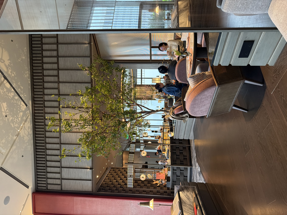 This image depicts a modern, upscale interior space, likely a hotel lobby or lounge, situated in Gongshu District, China. The environment features high ceilings with integrated recessed lighting and air vents, dark brown wooden plank flooring, and a sophisticated material palette including a prominent red vertical panel on the left, geometric patterned wall screens, and large windows on the right revealing an urban cityscape.
A large, multi-branched artificial tree with abundant green foliage serves as a central decorative element, extending towards the ceiling. Reflective glass or mirror panels on the right side integrate with the structural design, reflecting elements of the interior and exterior.
In the mid-right foreground, three adult individuals are seated around a circular table with a light pink surface. One person, wearing a light-colored sweater and glasses, faces left, appearing to be in conversation. Another, dressed in a dark top, is seated with their back partially towards the viewer. The third individual, wearing a blue jacket, faces right. Table settings include plates and a small vase.
To the background-left, a service counter or buffet display is visible. Two individuals, identifiable as staff by their dark attire, are positioned behind the counter, which displays various food items, pastries, and beverages. Notable decorative elements on the counter include large white rabbit figurines and circular, golden-hued light fixtures.
Seating arrangements include chairs with light pink velvet upholstery and wicker-style frames around the central table, and a grey upholstered sofa with a marble-topped side table and a brass-colored floor lamp on the far left. Other upholstered seating is visible throughout the lounge. Overhead and pendant lighting illuminates the entire scene.