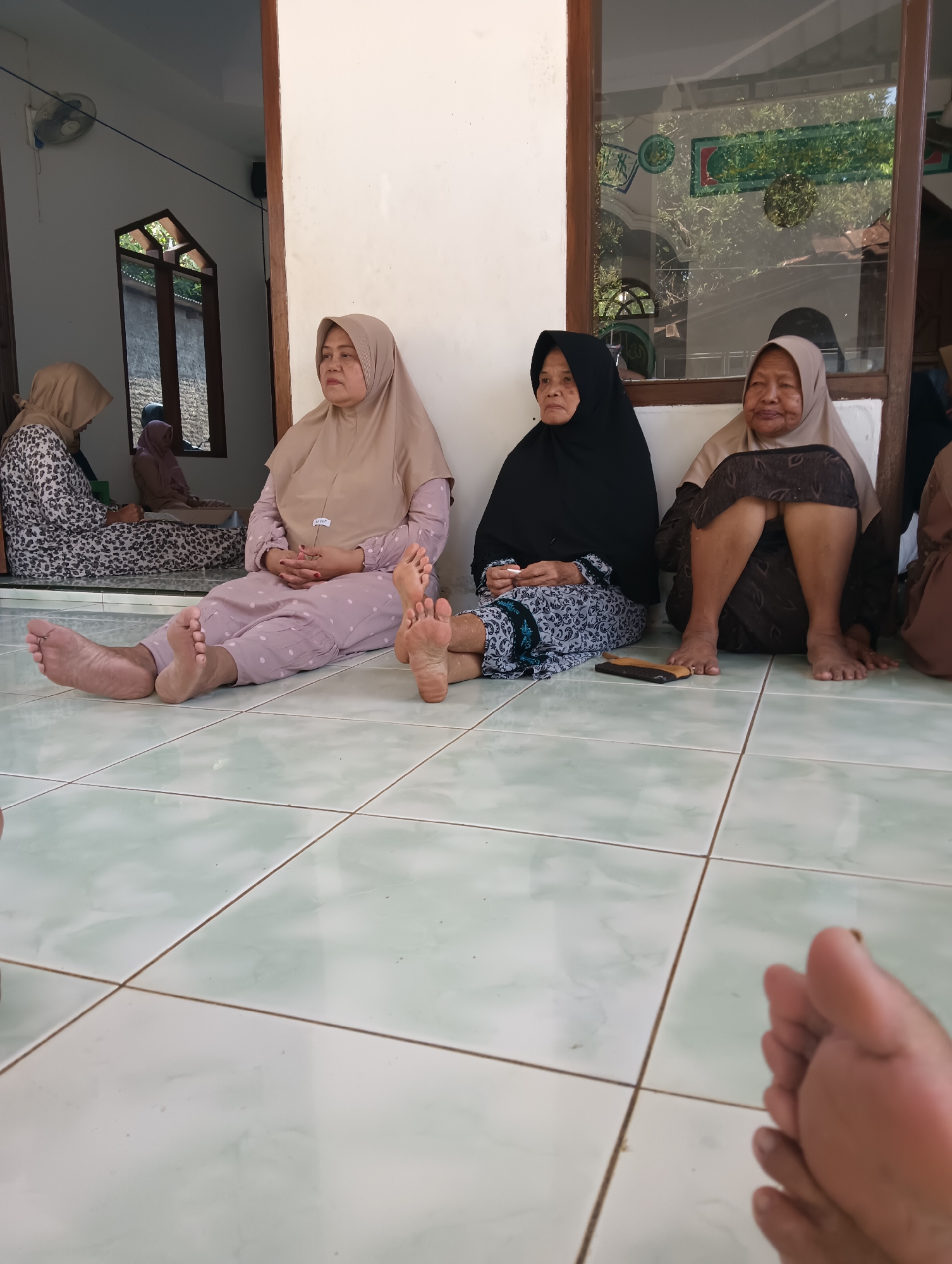 In Cluwak, Indonesia, four women are seated on a tiled floor, facing away from the camera. Three of the women are wearing headscarves and long dresses, while the fourth woman is wearing a black headscarf and a patterned dress. The woman in the foreground to the left is wearing a light brown headscarf and a pink dress with white polka dots, and her feet are visible. To her right, a woman in a black headscarf and a patterned dress is also seated, with her feet visible. Next to her, a woman in a light brown headscarf and a dark patterned skirt is seated with her legs bent. In the background, another woman in a light brown headscarf and a patterned dress is seated. The environment appears to be indoors, with a tiled floor and a white wall. A window with wooden frames is visible behind the women, reflecting trees and greenery. A fan is mounted on the wall in the background.