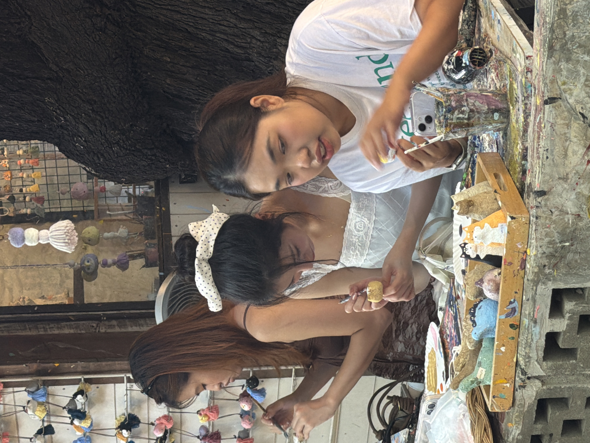 Three young women are engaged in a crafting activity at an outdoor table, possibly in Ban Sai Kham, Thailand, as indicated by the context. The woman on the left, with long brown hair, is focused on painting a small object held in her left hand, using a thin brush in her right. To her right, another woman with dark hair tied up with a white polka-dot bow is also painting a small, round object. The woman on the far right, wearing a white t-shirt with green lettering, is actively painting a small object, holding it with tweezers and a brush.
The table surface is heavily speckled with colorful paint, suggesting it is a dedicated workspace for art or crafts. Various art supplies are visible, including paintbrushes, a cup filled with brushes and a phone, a small hairdryer, and a wooden tray containing several painted objects, including cat figures and abstract shapes. Behind the women, a large tree trunk is prominent, and hanging decorations, possibly tassels, are visible against a backdrop that suggests an outdoor market or shop. The overall atmosphere is one of focused creativity and relaxed engagement in a shared artistic endeavor.