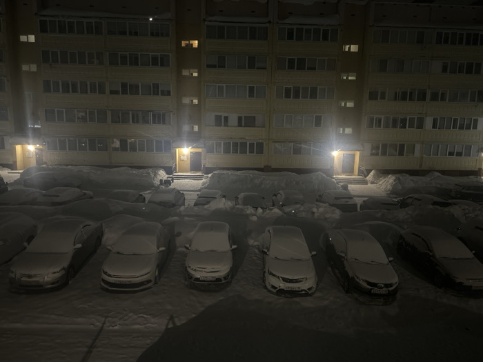 The scene depicts a snow-covered residential area at night in Noviy, Russia. Two multi-story apartment buildings, light yellow in color, dominate the background, featuring numerous windows, some of which are illuminated. Ground-level entrances, each with an overhead light, are visible between large snowdrifts piled against the building facades. In the foreground and midground, a parking lot is entirely covered in a layer of fresh snow. Multiple vehicles, approximately six distinctly visible in the immediate foreground, are parked and uniformly coated in snow, obscuring their details. The overall environment is dark, illuminated primarily by building lights and streetlights. No individuals or specific actions are observable.