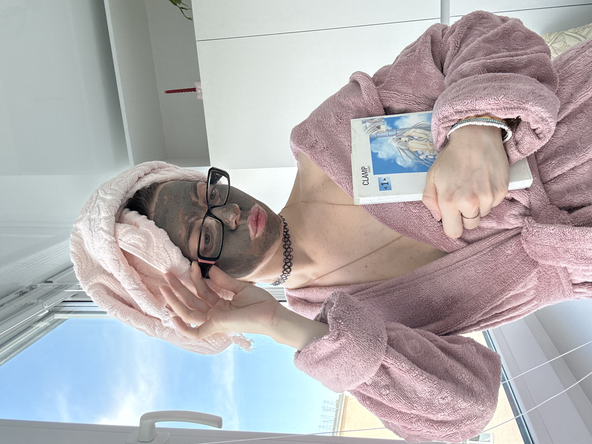 A young woman wearing a pink terrycloth robe and a pink towel wrapped around her head is applying a grey face mask. She has on glasses and a black choker necklace. She is holding a book with a picture of anime characters on the cover in her right hand. Her left hand is adjusted her glasses. She is standing in front of a window with a blue sky visible outside. There are shelves behind her with a red candle on them. The scene is indoors.