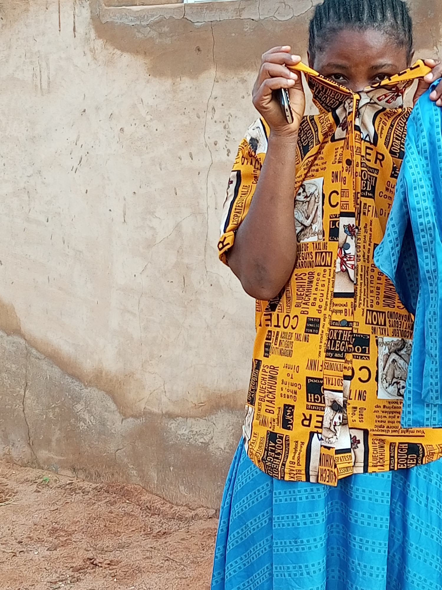 A medium shot captures a person in front of a textured, light brown, stucco wall. The person is wearing a yellow shirt with a black, white, and red text and image pattern, and a blue skirt with a geometric pattern. The person's hands are holding up the collar of the shirt to cover their lower face. A dark-colored mobile phone is held in the right hand, with the screen facing the viewer. The person's eyes are visible above the shirt. The wall behind the person shows signs of wear, with visible cracks and patches of discoloration. The ground in the foreground is a reddish-brown, sandy soil. The overall lighting suggests an outdoor setting. The image does not provide any specific visual cues to pinpoint a location within Tempure, Nigeria.