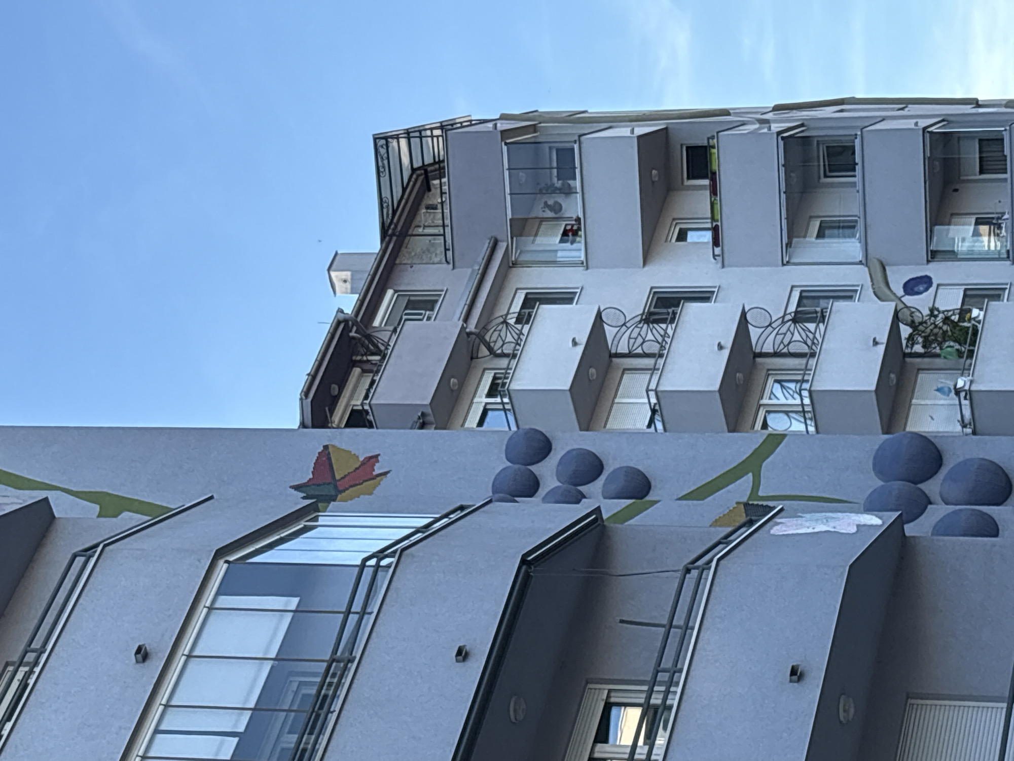 The image shows two buildings under a clear blue sky in Kragujevac, Serbia. The lower building is light gray with a mural depicting stylized flowers and green stems, as well as blue hemispherical shapes. It has several sloping windows with dark frames. A metal ladder runs vertically up the side of one of the architectural projections.
The taller building behind it is also light gray and appears to be an apartment building with projecting box-like balconies. Some balconies have ornate metal railings, and some appear to be enclosed with glass. Windows are visible on each level.