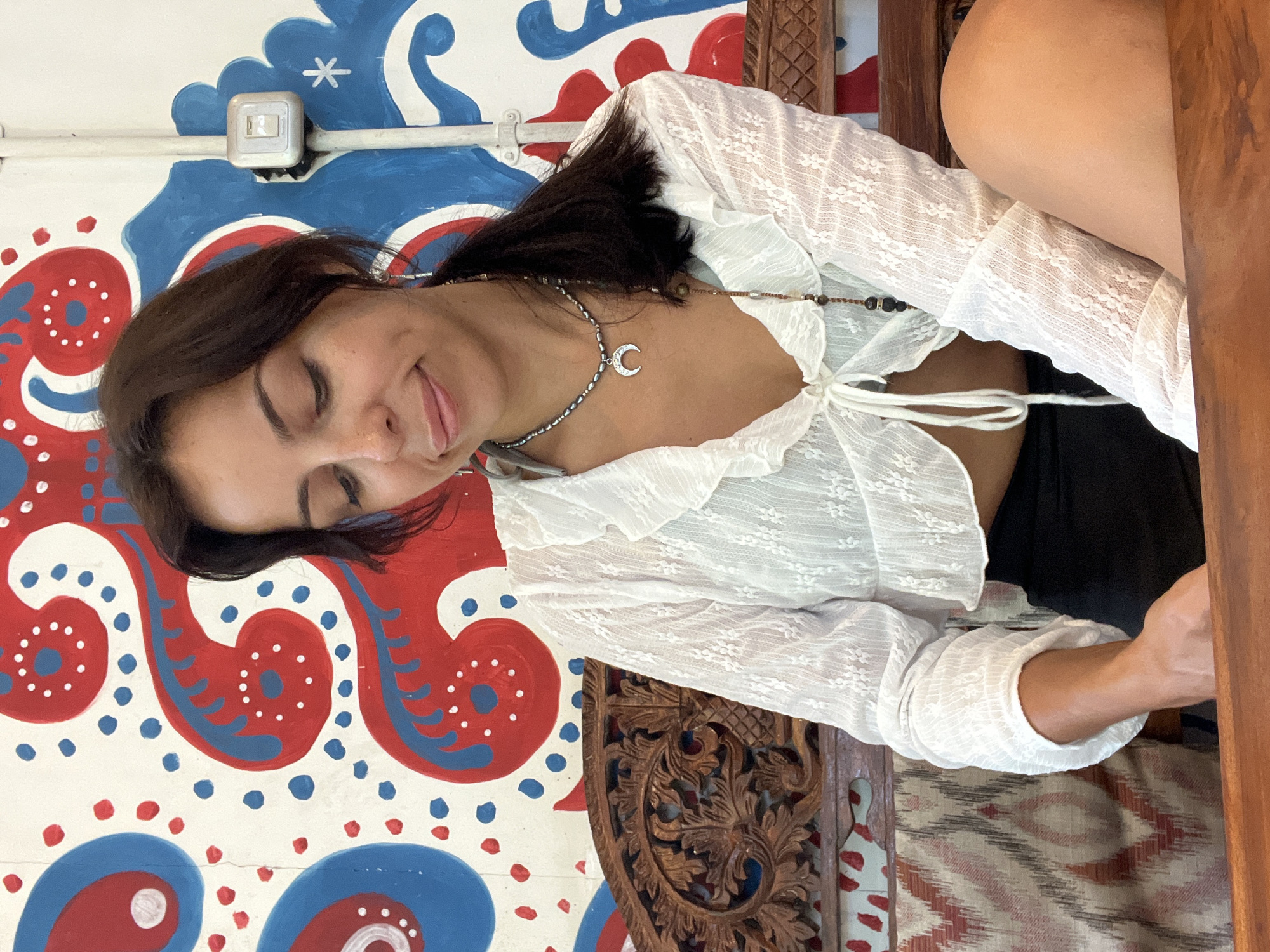 A vibrant, daytime portrait features a woman captured from the chest up, looking upward and slightly to her left. She has dark hair and wears a white, sheer, lace long-sleeved crop top with ruffled edges and a tie-front closure. A silver necklace with a crescent moon pendant adorns her neck, along with a secondary beaded necklace. Her eyes are closed, and a gentle smile plays on her lips, with her tongue peeking out, conveying a sense of playfulness or peacefulness.
The background is a white wall decorated with a bold, hand-painted pattern in red and blue, featuring large swirling designs accented with white and blue dots. A white electrical light switch is visible on the wall in the upper left of the frame. To the woman's right, and partially obscuring the foreground, is an intricately carved wooden furniture piece, possibly a chair or decorative panel, displaying elaborate floral or geometric patterns. A patterned fabric, resembling a rug or upholstery with a chevron-like design in muted colors, is also visible in the lower right. The natural lighting and the woman's light clothing suggest a warm climate. The colorful wall art and carved wood hint at a bohemian or artisanal setting, aligning with the location context of Kuta Selatan, Indonesia. No visible text is present.