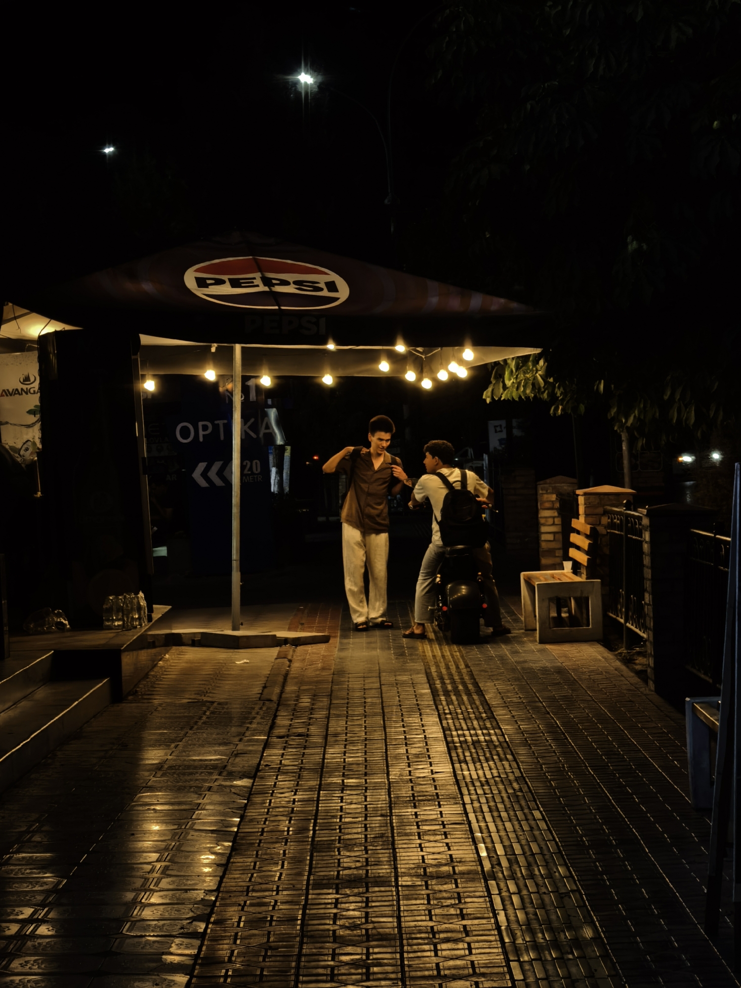 The image depicts an outdoor scene at night, possibly in Fergana City, Uzbekistan, under artificial lighting. Two young men are present. One man, wearing a brown shirt and light-colored pants, stands with his left arm bent and hand on his hip, facing towards the right of the frame. The other man is seated on a dark-colored motorcycle, facing left and looking at the first man. He is wearing a light-colored t-shirt and pants, and has a dark backpack on. They appear to be interacting or conversing.
Above them, a large umbrella with the "PEPSI" logo is visible, illuminated by strings of bare light bulbs. Behind the men, there is a structure with signage that includes the word "OPTKA" and some text that appears to be in a Latin-based alphabet, potentially indicating a business. To the left, there is a dark structure with some bottles on a step. The ground in the foreground is a patterned walkway made of rectangular tiles, reflecting the light. To the right, a metal fence and brick structures are visible. The overall atmosphere is dimly lit, with strong highlights and shadows contributing to the nighttime setting.