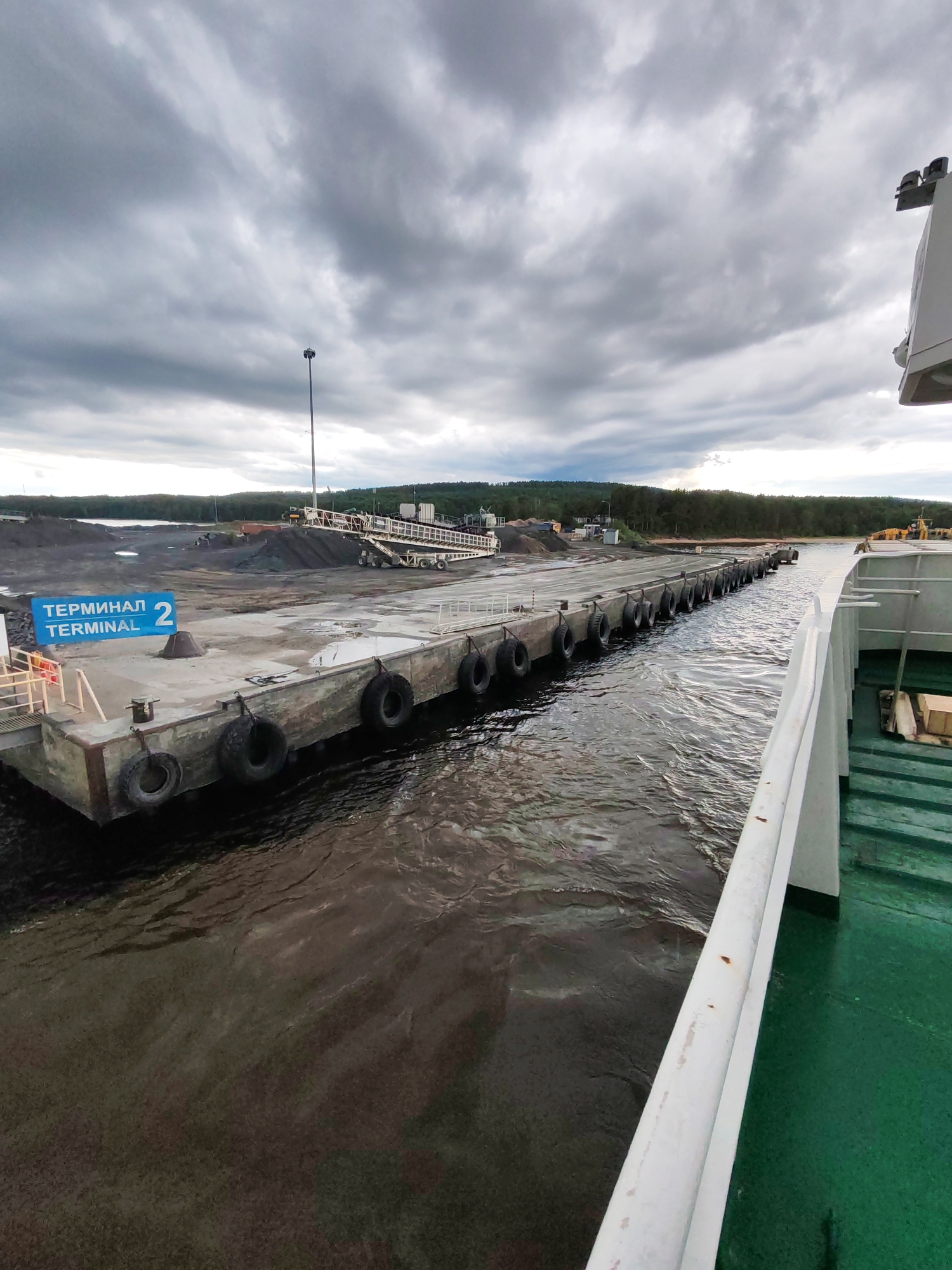 The image shows a dock at Terminal 2 in Shcheleiki, Russia. The dock is made of concrete and is lined with tires to protect ships. There is a ramp leading from the dock to the shore, where piles of dark material are visible. The sky is overcast with heavy clouds. Part of a green-decked vessel is in the foreground, on the right side of the image.