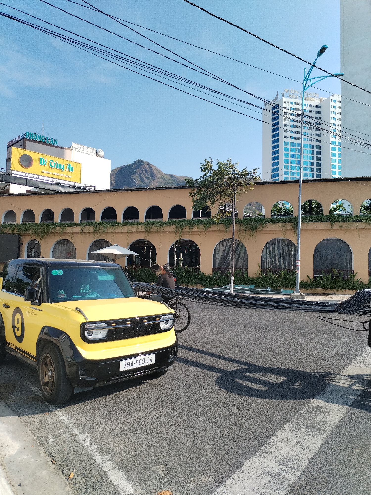 The image captures a street scene in Bac Nha Trang Ward, Vietnam. A distinctive yellow and black VinFast VF5 electric vehicle with the number "9" prominently displayed on its side is parked on the left side of the road. The license plate reads "79A-569.04". In the background, an arcaded building with tan-colored walls and green vines hanging from the arches stretches across the midground. Above this building, a sign reads "PHONG LAN" and "Dê Tràng An". Behind the arcaded structure, a distant mountain range is visible. To the right, a tall, modern building with blue windows contrasts against the clear blue sky. A turquoise street light pole stands nearby. A person on a bicycle is visible near the center of the image, partially obscured by the VinFast and the nearby objects. Metal rods and a bicycle wheel cast shadows on the pavement.