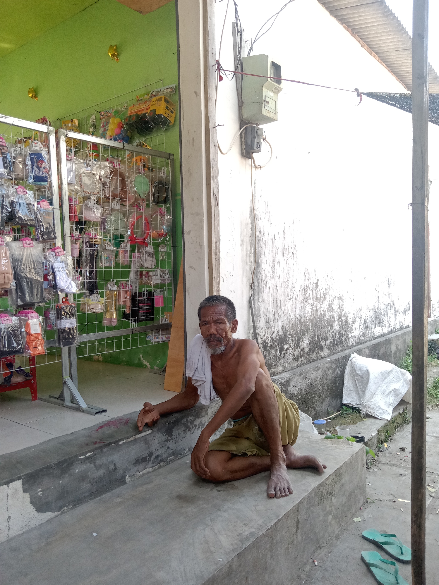 The image depicts an outdoor scene in Klepek, Indonesia, likely in front of a small shop. A man, appearing elderly and shirtless, sits on a concrete ledge. He is wearing light-colored shorts and has a white cloth draped around his neck and shoulder. His gaze is directed towards the camera.

To the left of the man, a shop displays various items on metal racks. These items appear to be clothing or accessories, packaged individually and hanging from the shelves. The shop's interior wall is painted bright green, and a yellow toy truck is visible on a shelf.

To the right of the man, a white wall with visible water stains and grime forms the backdrop. Electrical equipment, including a meter box, is mounted on this wall. A white, loosely tied bag rests on the ground near the wall. The ground in front of the ledge is concrete, with a pair of green flip-flops visible in the lower right corner. The overall environment suggests a modest, possibly semi-rural setting.