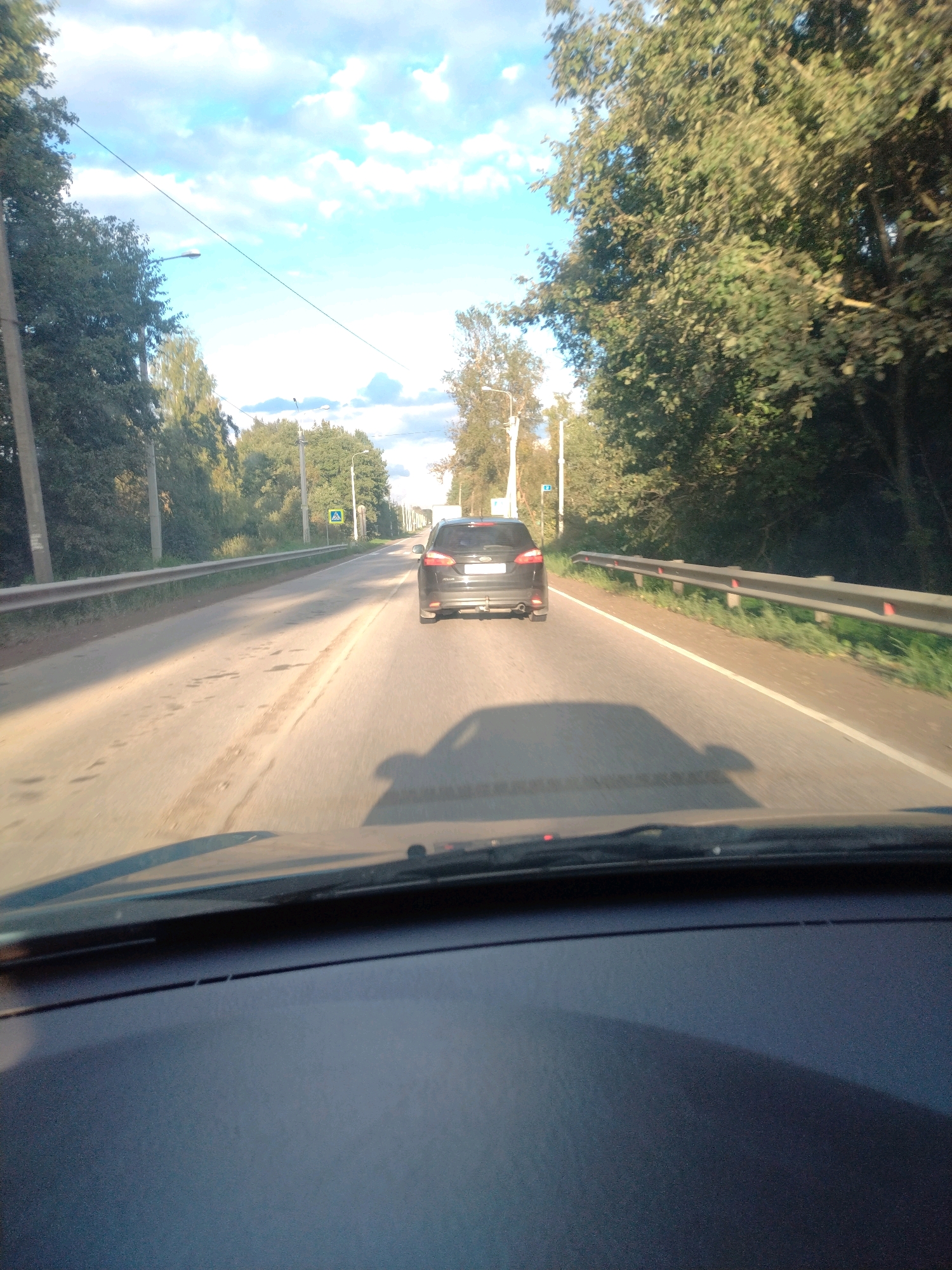 The image shows a view from the driver's seat of a car on a paved road. The car is moving forward. The road is bordered by trees and grass on both sides. A black car is visible ahead, also moving in the same direction. There is a pedestrian crossing sign on the right side of the road. The sky is blue with some clouds. The location is identified as Krasnoborskoye, Russia.