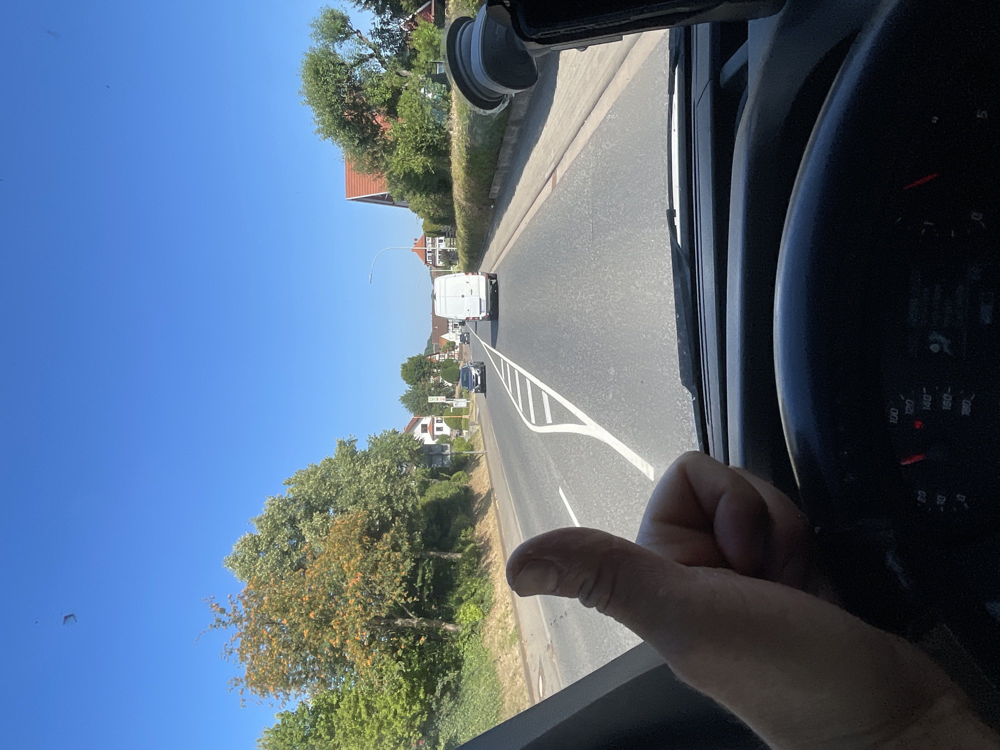An outdoor, eye-level, forward-facing shot captures a driver's hand giving a thumbs-up from inside a vehicle. The driver's thumb is prominently positioned in the foreground, and a portion of the steering wheel and dashboard gauges are visible at the bottom of the frame. The vehicle is on a paved road in what appears to be a suburban area. A white cargo van is driving ahead on the road, followed by a dark-colored car. The road is lined with trees and houses with red-tiled roofs on either side. The sky is clear and blue, indicating good weather. The scene is likely in Wartenberg, Germany, given the architecture and the style of the vehicles.