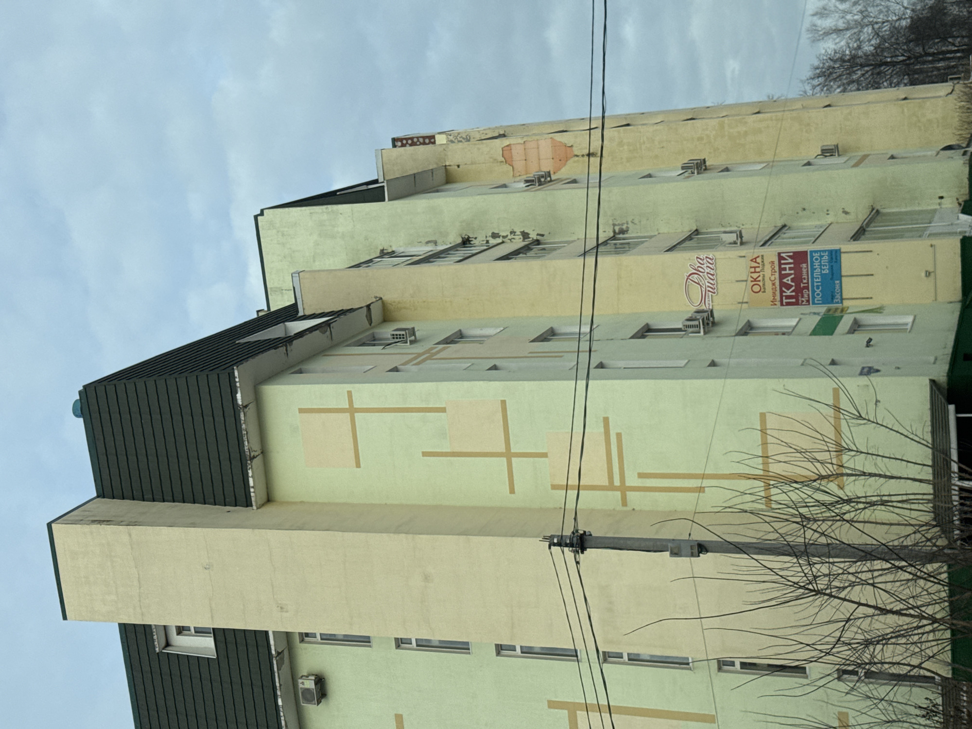 The image depicts an exterior view of a multi-story building in Chaikovsky, Russia. The building's facade is primarily light green and yellow, with painted geometric patterns on some sections. It has multiple windows and several air conditioning units are visible. Signs in Russian, advertising "Okna" (Windows), "Tkani" (Fabrics), and "Postel'noe Bel'e" (Bed Linen), are affixed to the building. The top of the building has a dark green roof. Electrical wires are strung across the front of the building. Bare tree branches are visible on the right side of the frame, obscuring part of the building. The sky is overcast.
