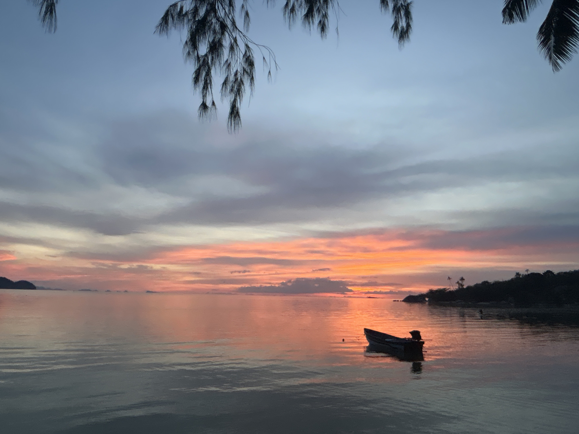 This image captures a serene sunset scene on Koh Phangan island, Thailand, specifically near the area of Tambon Ko Pha Ngan. The sky is a canvas of soft blues, purples, and vibrant oranges, transitioning into pink hues as the sun dips below the horizon. Wispy clouds are scattered across the sky, reflecting the warm colors of the sunset. In the foreground, calm water stretches towards a silhouetted coastline dotted with trees. A small, dark boat with an outboard motor is moored in the water, its reflection shimmering on the surface. Tropical foliage hangs into the frame from the top, adding a natural border to the tranquil view. The overall impression is one of peacefulness and natural beauty.