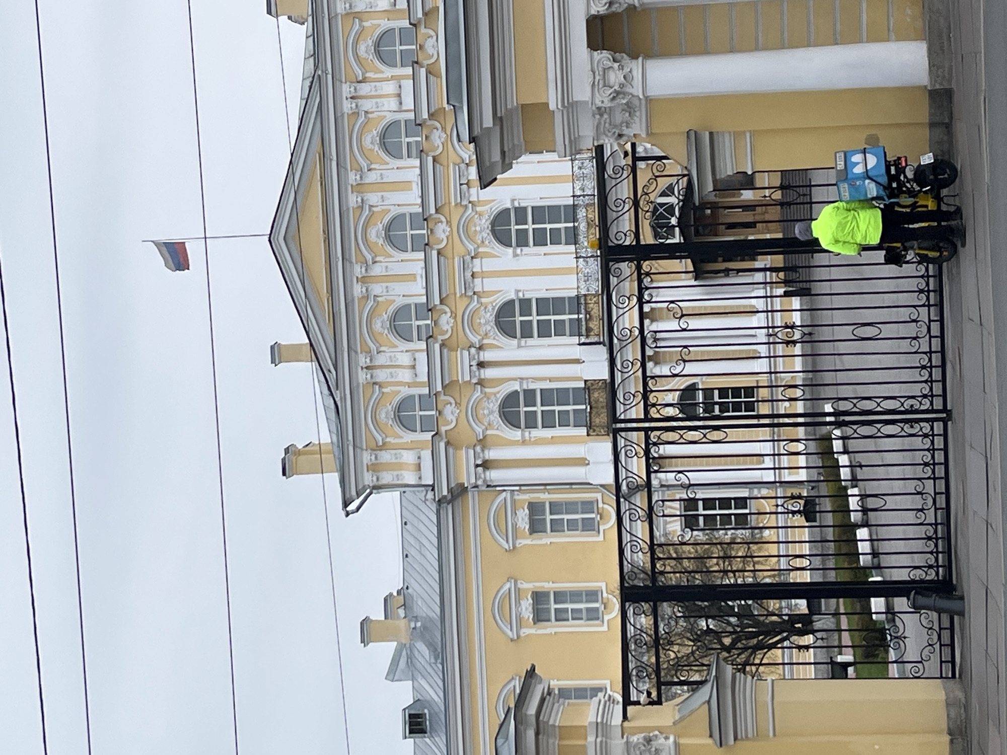 The image shows a portion of a yellow and white building with ornate architectural details, viewed through a black iron gate. The building has multiple windows and decorative elements, including a flag on the roof. A person in a bright yellow jacket is near a bicycle with what appears to be delivery boxes attached, positioned near the gate. The sky is overcast. The location appears to be urban.