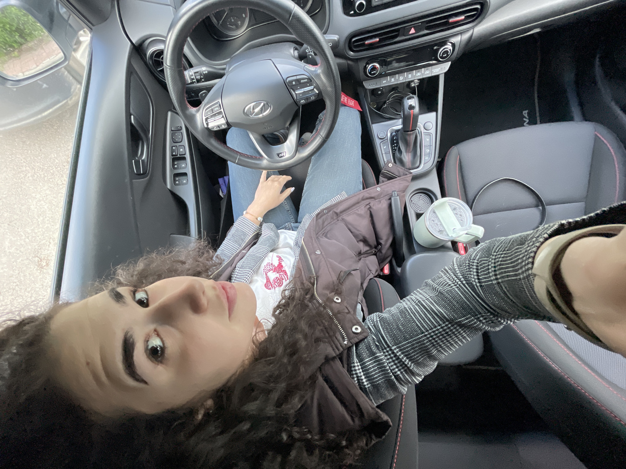 A woman with curly dark hair is seated in the driver's seat of a Hyundai Kona. She wears blue jeans, a white t-shirt with a red design, a plaid shirt-jacket, and a dark brown vest. Her left hand rests on the steering wheel. A cup is in the center console cupholder. The interior of the car has dark grey seats with red accents. The image provides no contextual information to determine its location in Sofia, Bulgaria.
