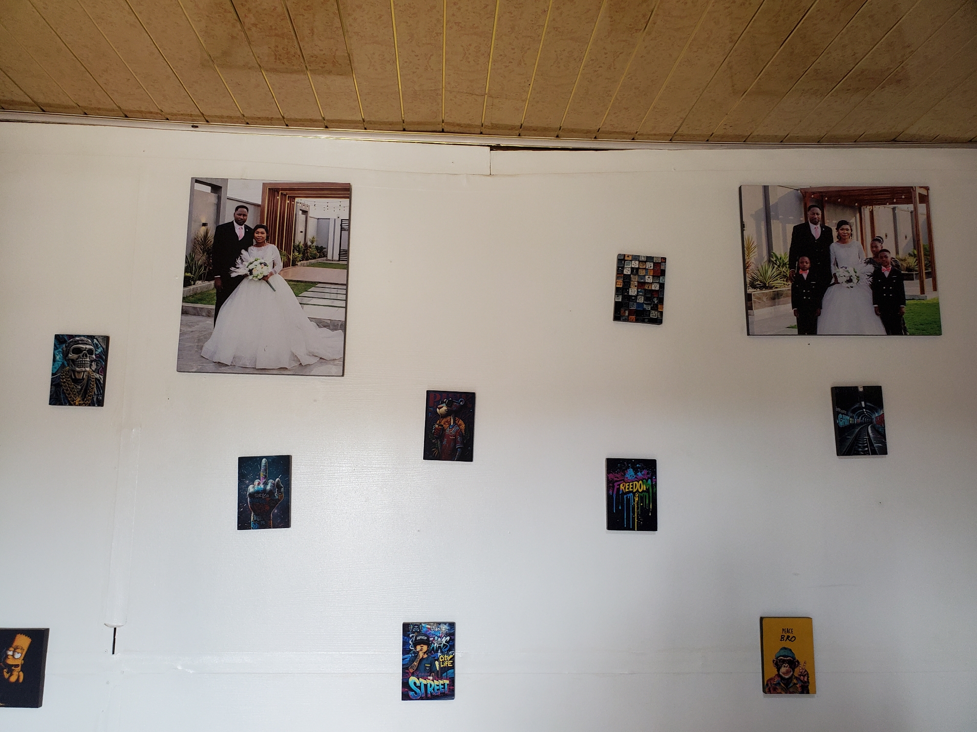 The image shows a wall decorated with several pictures. At the top of the wall, two larger wedding photos feature a bride in a white gown and a groom in a suit. One photo shows the couple alone, while the other includes two children. Below the wedding photos are smaller pictures, including a skull, a hand with a raised middle finger, and images with the words "Freedom" and "Street". One picture appears to feature Bart Simpson. The wall is white, and the ceiling is light brown. It is not possible to determine the location in Jos, Nigeria from this image alone.