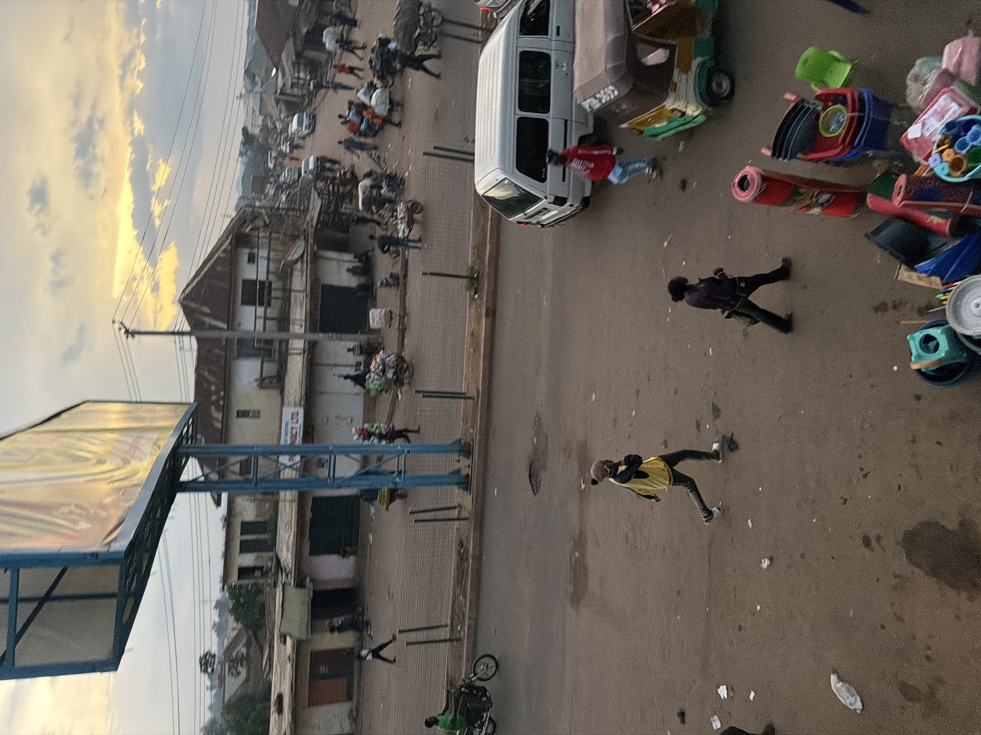 The image depicts a street scene in Bukuru, Nigeria, with a street market atmosphere. The time of day appears to be late afternoon or early evening, as indicated by the warm lighting and cloudy sky. The foreground shows a dusty road with scattered debris and a few people.
A person in a yellow and black jacket with dark pants and white sneakers is running across the road. To their left, another person in a dark jacket and dark pants is walking. In the background, there are several buildings, some appearing to be shops or market stalls. A white van is parked on the right side of the road. Further down the street, more vehicles and people are visible, along with market stalls laden with goods. A man on a green motorcycle is seen on the left side of the road. Overhead, a large billboard structure with an advertisement is visible. The overall impression is of a busy urban environment with elements of commerce and daily life.