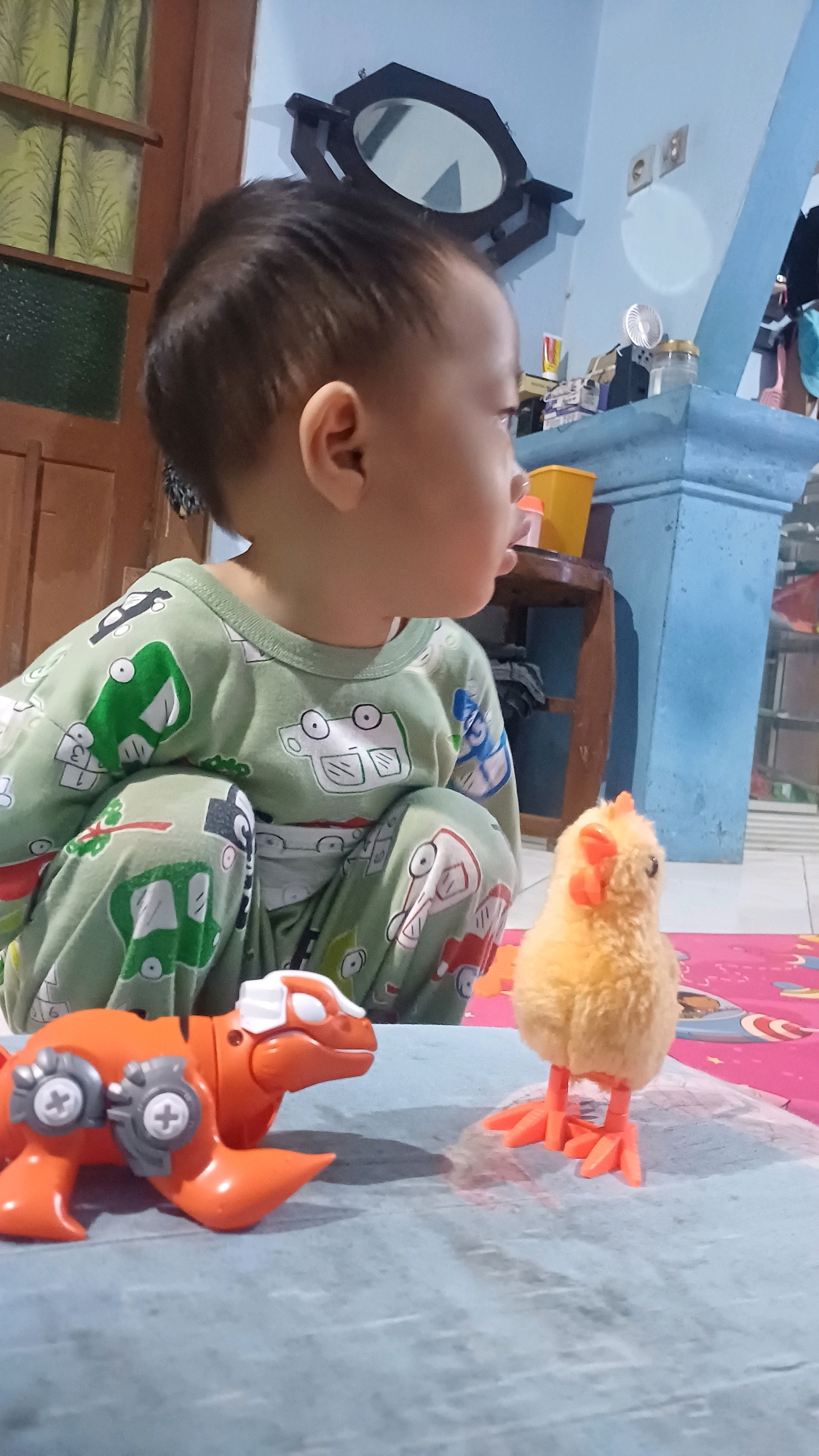 In a domestic setting in Wonokerso, Indonesia, a young child, likely a toddler, is seated on the floor, wearing a light green pajama set with a pattern of cars. The child is looking to the right. In front of the child are two toys: an orange dinosaur toy with grey mechanical details and a small, fluffy yellow chick toy with orange feet. The background reveals a blue wall with a mirror hanging on it and a wooden cabinet. The floor appears to be tiled and covered in part by a pink mat. The lighting suggests an indoor environment during the day.