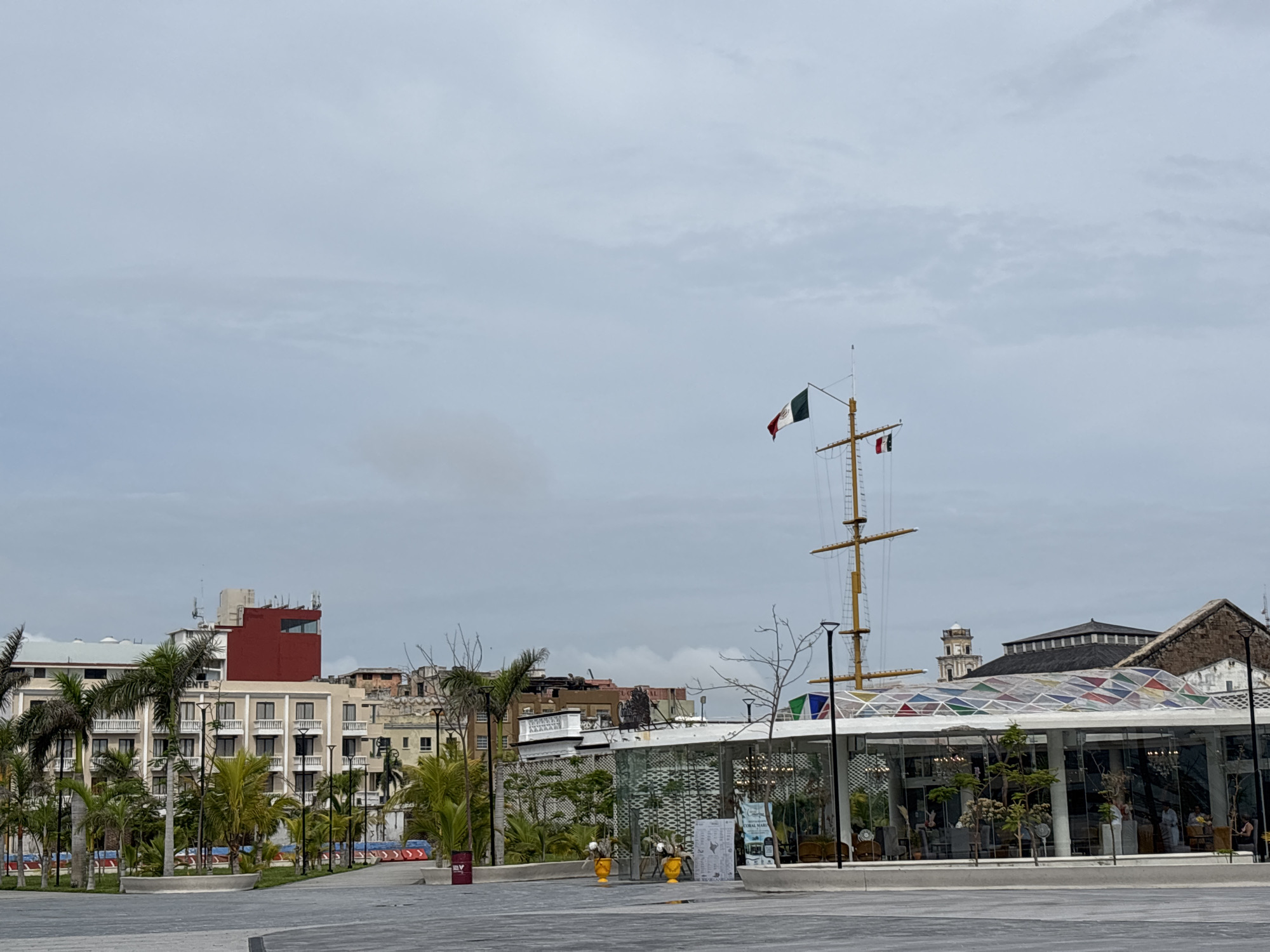 The image captures a view of the Malecon in Veracruz City, Mexico, on an overcast day. In the foreground, a paved open area leads to a modern building with a glass facade and a colorful, multi-paneled roof. This building appears to be a cafe or restaurant, with tables and chairs visible inside. Adjacent to this building is a tall mast with several flags, including the Mexican flag, hoisted on a ship-like structure.
Behind the modern building, a multi-story hotel with balconies and palm trees in front of it is visible. Further in the background, various other buildings of different architectural styles and heights dot the skyline, some showing signs of age. The sky is uniformly cloudy, casting a soft light on the scene. No people are clearly discernible in the image.