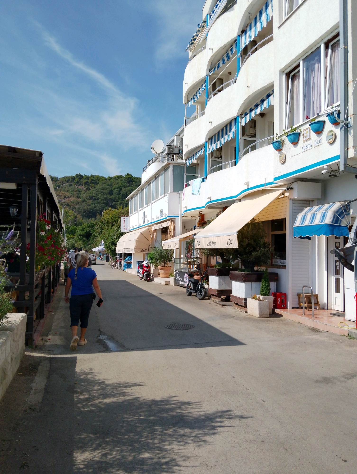 A woman in a blue shirt and dark capri pants walks away from the camera down a paved street in Sutomore, Montenegro. She is holding a dark object in her right hand. To her left is a covered walkway with wooden posts, adorned with hanging plants, including red and white flowers. To her right is a white, multi-story building with blue and white striped awnings and balconies. The building has signs indicating "VISTA ORO" and other businesses. The street is lined with businesses and planters. In the background, a lush green hillside rises under a blue sky with sparse white clouds. The sunlight casts shadows on the street.