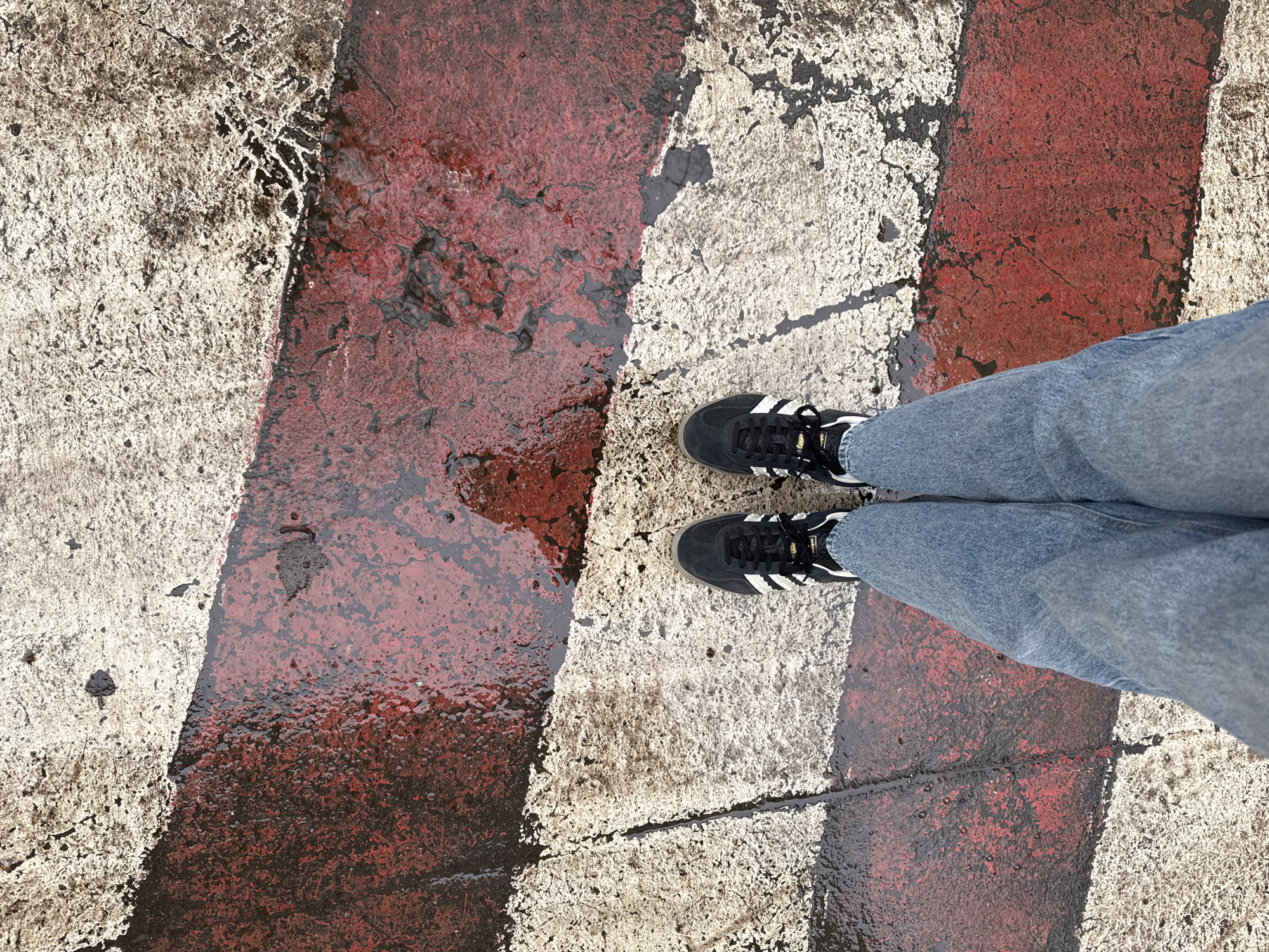 A person is standing on a crosswalk, with their feet positioned on two adjacent stripes. The crosswalk has alternating stripes of red and white. The surface appears wet, possibly from rain, reflecting the colors of the stripes. The person is wearing blue jeans and black Adidas sneakers with white stripes. The setting appears to be in an urban environment, possibly Moscow, Russia, based on the architectural style and the color scheme commonly found in crosswalks there. The image is taken from a high angle, looking down at the subject.