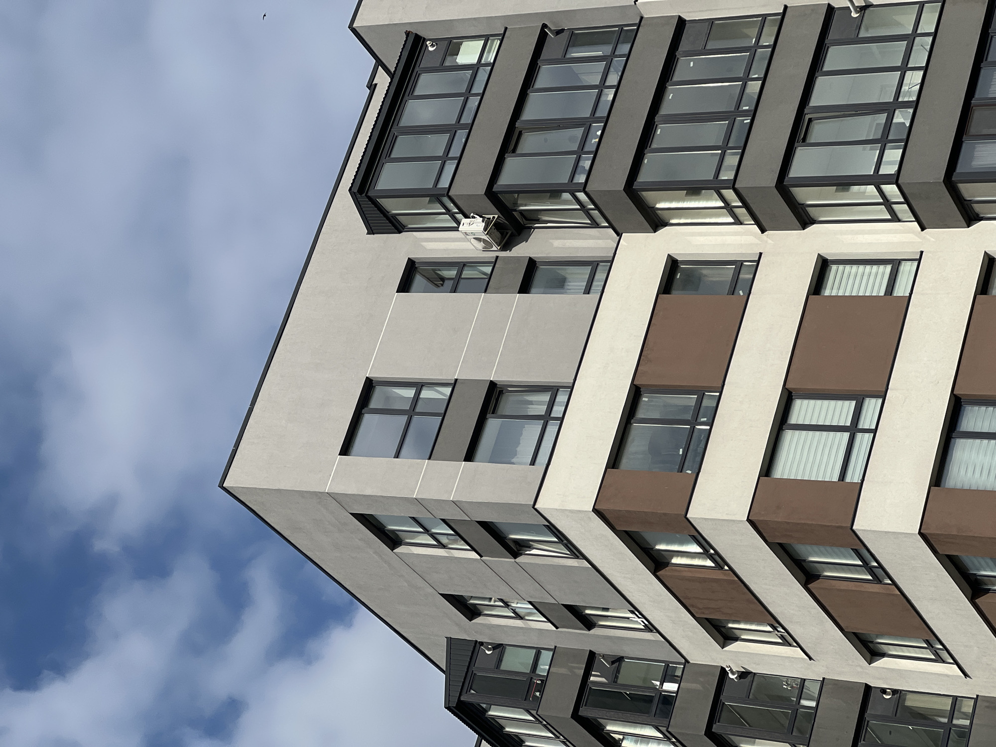 A low-angle, full shot captures the exterior of a modern apartment building against a bright blue sky dotted with wispy white clouds. The building exhibits a contemporary architectural style with distinct geometric sections and varying color palettes. The visible facades are primarily a light grey, with prominent horizontal bands of dark brown and off-white. Large, dark-framed windows are a dominant feature, with some featuring horizontal blinds. A section of the building features a cantilevered, multi-story glass enclosure with dark frames, likely a balcony or sunroom. An outdoor air conditioning unit is mounted on the grey facade near this glass enclosure. The overall impression is one of a new, urban residential structure.