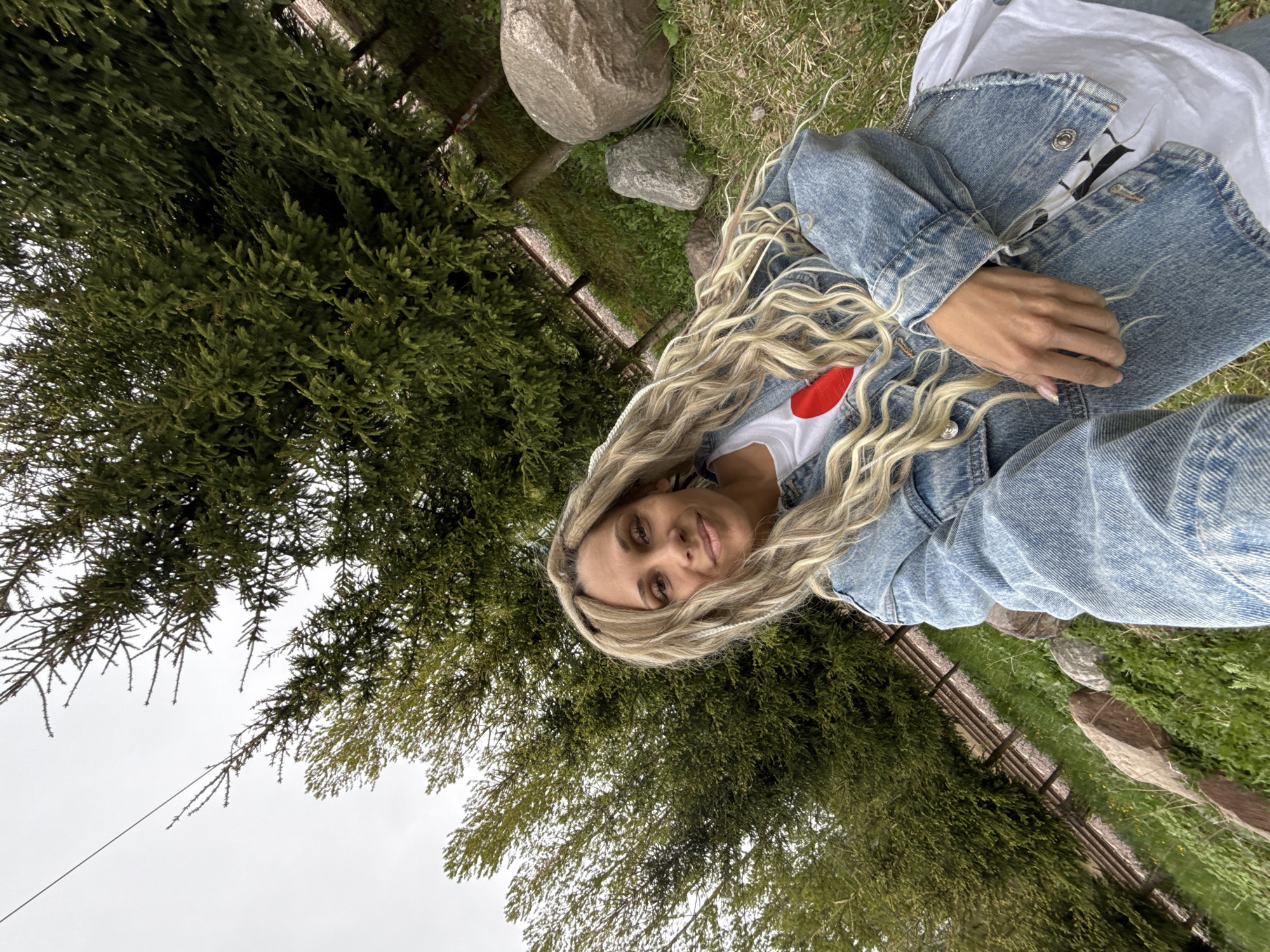 A woman is lying in a grassy area in Шебунино, Russia. She is wearing a denim jacket over a white t-shirt with a red heart. She has long, blonde wavy hair. A large evergreen tree is nearby, and a fence, grass, and rocks are in the background. The sky is overcast.