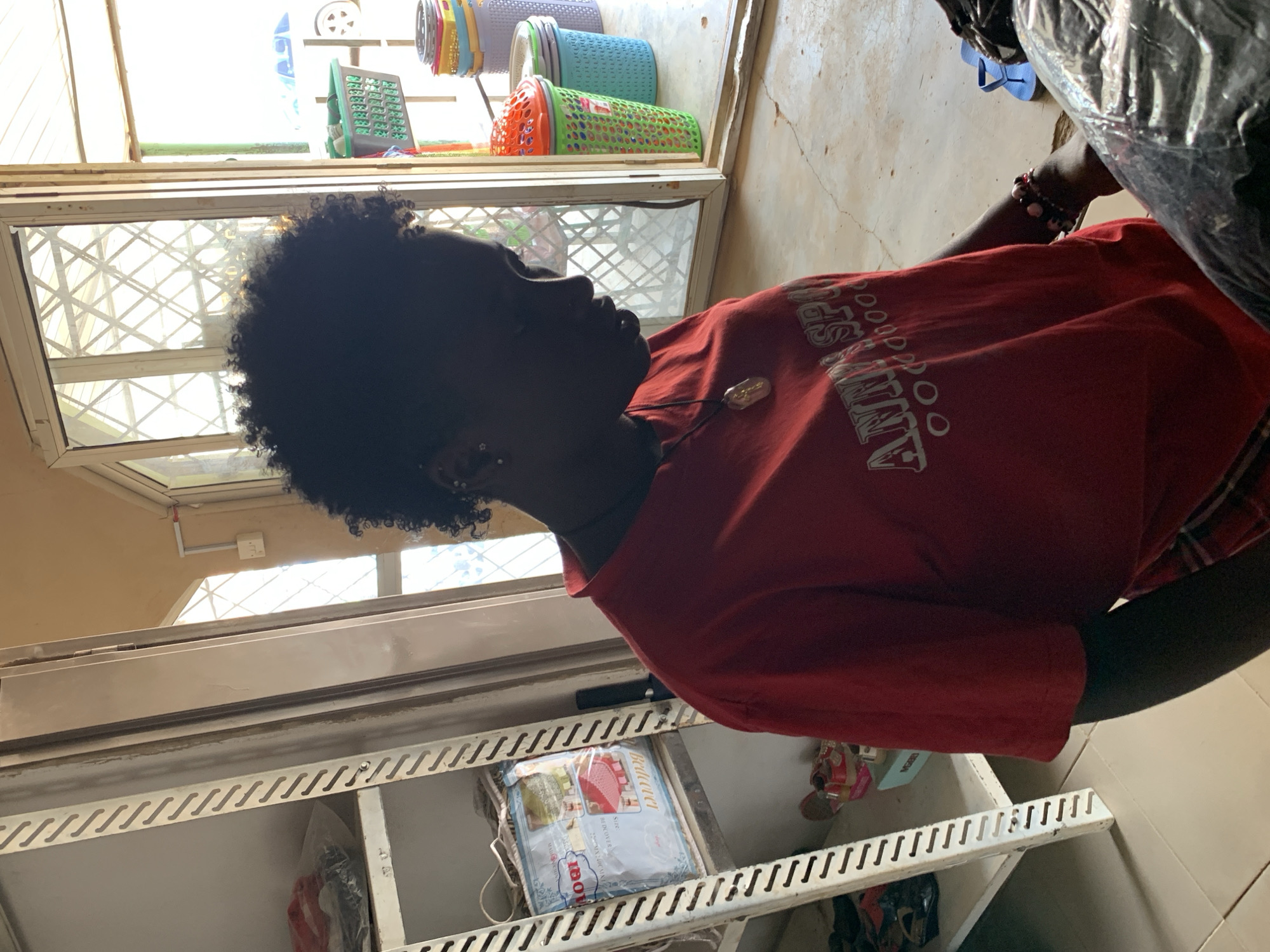 A young Black female wearing a red t-shirt with white text that reads "ANN SP" and a black leggings is standing in what appears to be a shop or storeroom. The female is silhouetted against a bright background, obscuring details of her face, but her curly dark hair is visible. She is wearing a necklace with a pendant. To her left is a white metal shelving unit filled with various items, including packaged goods and what appears to be clothing. To her right, through a doorway or window, several colorful plastic baskets are stacked. The floor is concrete and appears somewhat dusty. A blue flip-flop is visible on the floor near the doorway. The scene is lit by natural light coming from the right side of the image, suggesting it is daytime. The overall impression is of a casual, domestic, or small business environment.