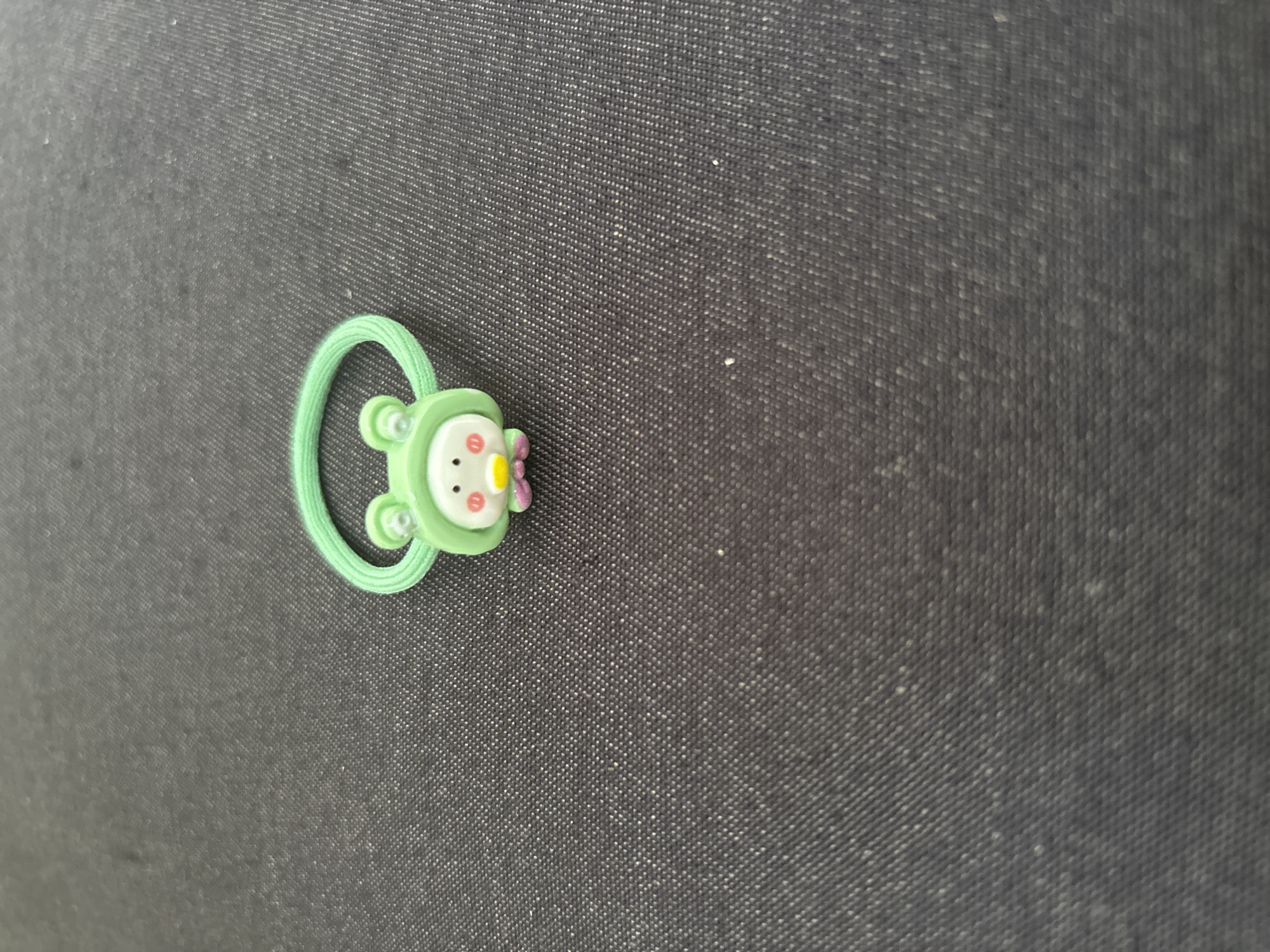 A cute, green frog-shaped hair tie is centered on a dark, textured surface. The frog has large, round eyes, a small yellow beak, and pink cheeks. Its body is green and appears to be made of soft material, possibly rubber or silicone. The hair tie itself is a thin, bright green elastic band. The background is a dark, likely fabric, surface with a subtle woven pattern. There are no people or any other objects in the image. The scene is a close-up shot, with the hair tie being the sole focus. There is no identifiable information that suggests a location in the city of Imeni Morozova, Russia.