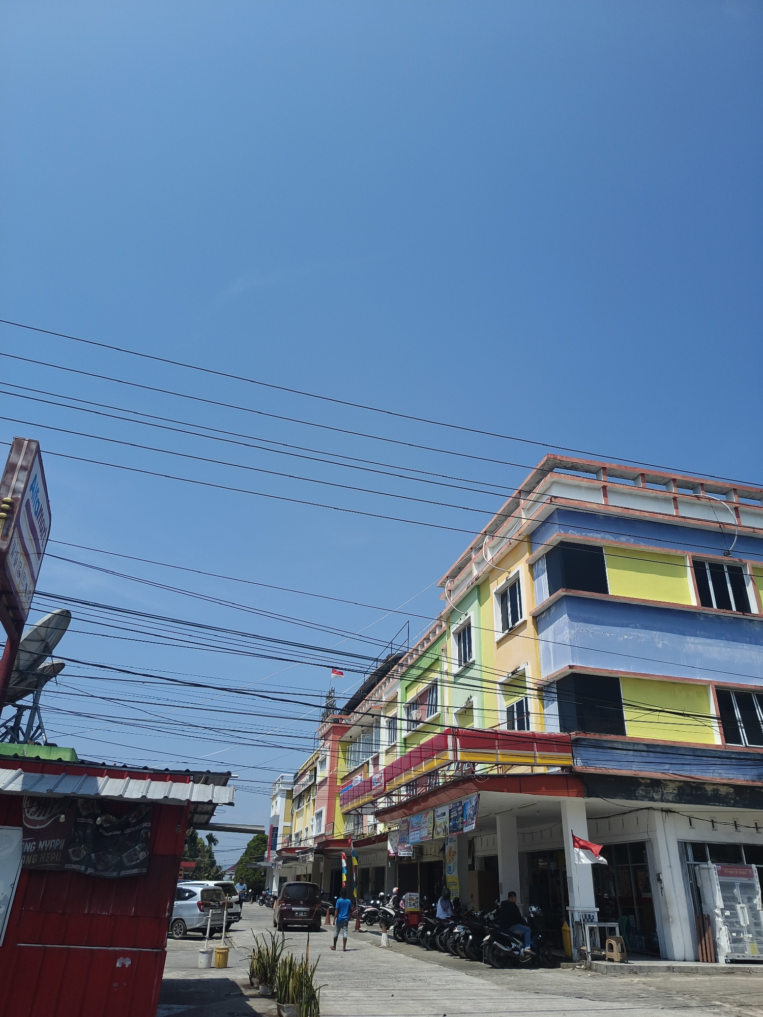 This image depicts a street scene in Pangkalpinang, Indonesia, under a clear blue sky. The foreground features a red roadside stall with a corrugated metal roof and a sign. To the right, a multi-story building with a vibrant facade, painted in stripes of blue, yellow, and green, dominates the view. The building has multiple windows and balconies with red trim. Several electrical wires are strung across the sky, obscuring parts of the buildings and the sky. Parked motorcycles are visible along the sidewalk in front of the colorful building. A few individuals are present on the street. One person in a blue shirt walks away from the camera, while others are seated on motorcycles or walking in the background. Cars are also parked on the street. The overall impression is a busy urban environment, likely during daylight hours.