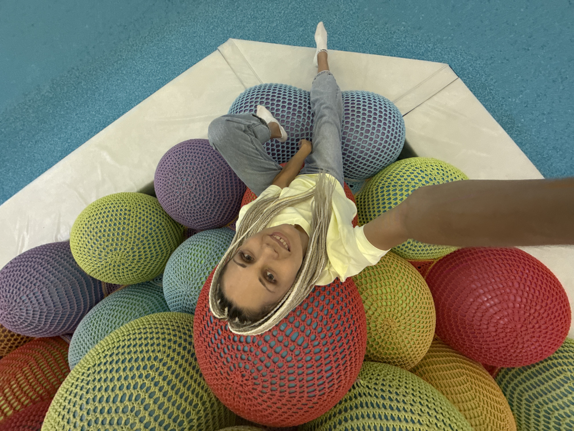 The image shows a woman lying on a pile of large, colorful crocheted balls. The balls are red, yellow, green, blue, and purple. The woman is wearing a yellow t-shirt and blue jeans. She has blonde braided hair and is smiling at the camera. The background is a bright blue surface, possibly a pool or a soft play area. This scene is not identifiable as being in Korobovo, Russia, or any specific city.