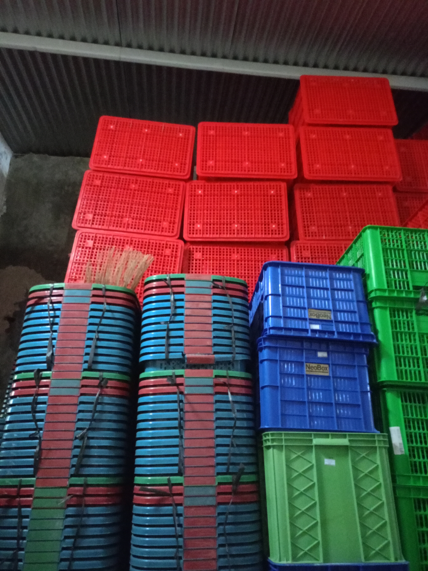 In a dimly lit warehouse in Batu, Indonesia, a large quantity of plastic crates are stacked high. Red crates are piled in the upper sections of the image, some open and some stacked, revealing a mesh-like pattern. Below these, a collection of vertically stacked crates in red, blue, and green are visible, secured by thin ties. A cluster of brooms or brushes protrudes from the top of one stack. In the lower right, two large blue crates with "NeoBox" branding are stacked atop each other, followed by a bright green crate. Further to the right, several large green crates, also with a mesh design, are stacked. The environment suggests a storage or distribution facility, with the corrugated metal ceiling of the warehouse visible overhead. There are no people or animals present in the image.