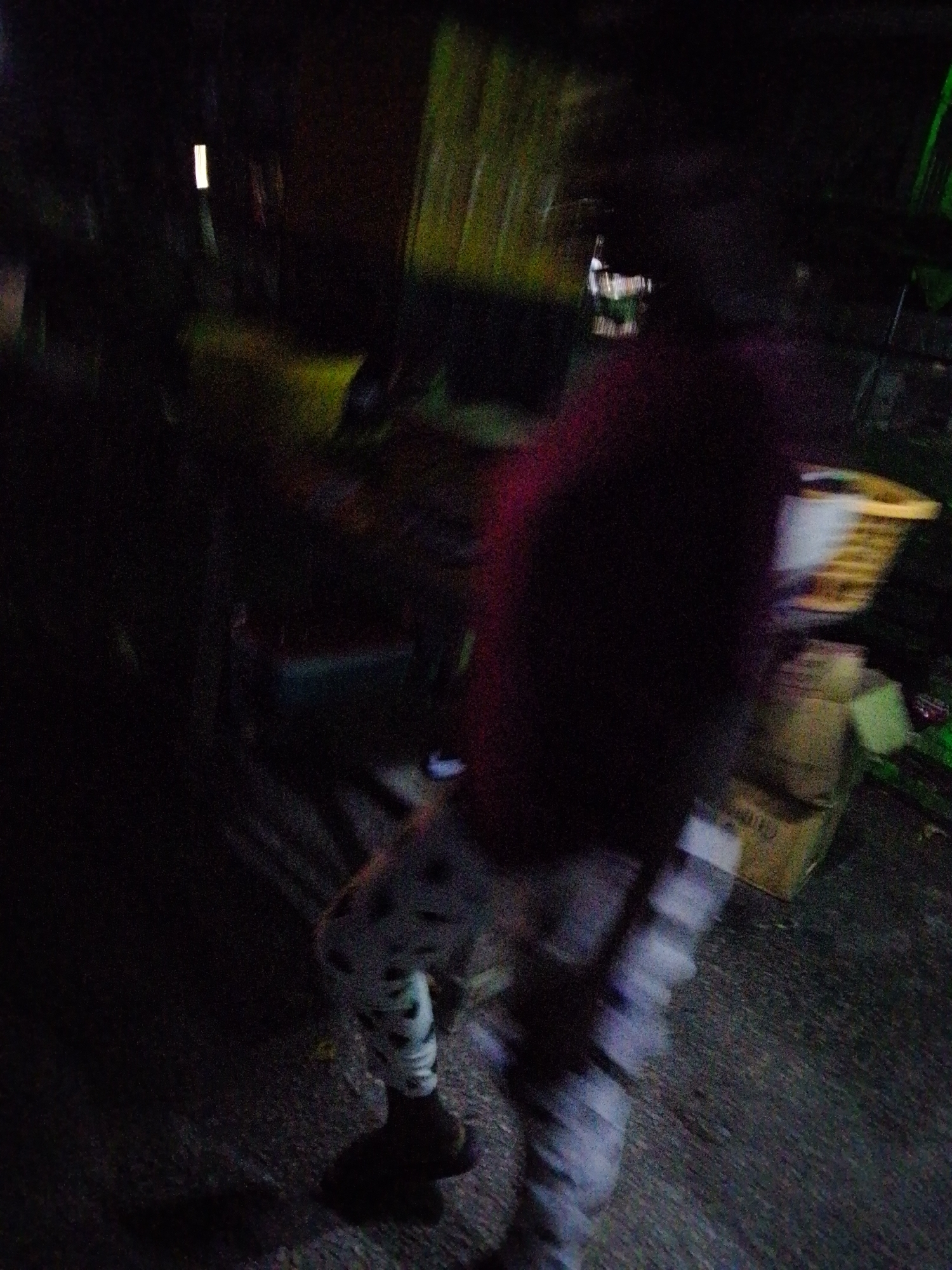 The image depicts a blurry, dark scene that appears to be an outdoor area at night. A person is visible, their form blurred due to motion or low light conditions. They are wearing pants with a patterned design, possibly black and white stripes or checks, and dark shoes. The person is standing in what seems to be an unpaved or rough surface, possibly a dirt or gravel ground.
In the background, there are out-of-focus objects that suggest a storage or backlot area. To the right, there are cardboard boxes stacked, one of which has some visible markings. A yellow object with horizontal slats, possibly a basket or a rack, is situated above the boxes. The overall environment is dimly lit, with some faint streaks of green light visible in the upper left and right portions of the image, possibly from distant lights or foliage. There is a distinct vertical white light source in the upper left, also out of focus.
The context of Jalingo, Nigeria, suggests this scene could be a part of the city's commercial or residential back areas, perhaps a market stall, a residential compound, or a less developed urban space. The image captures a moment in time characterized by low visibility and obscured details.