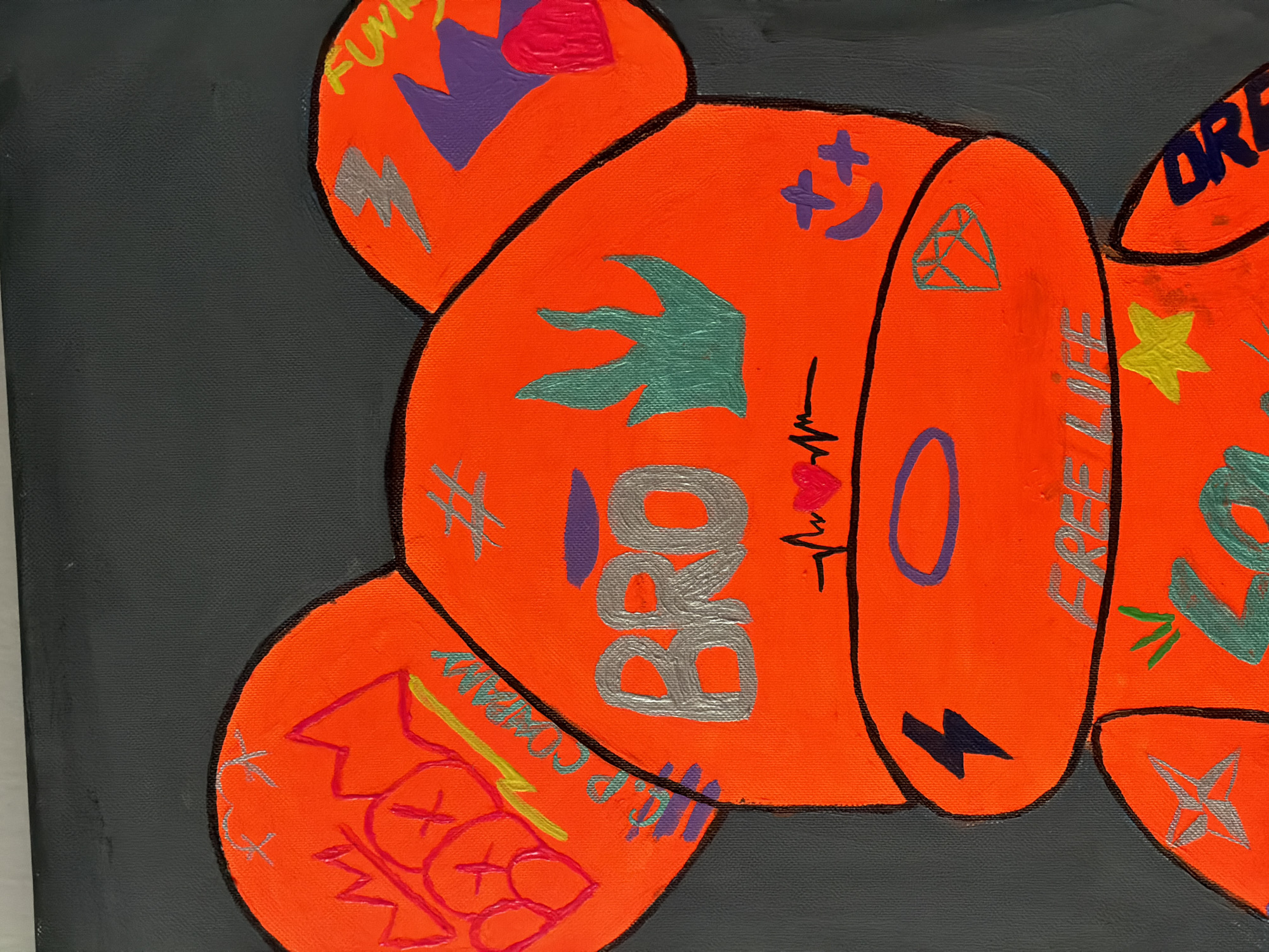 The image shows a close-up of a painting of a stylized bear figure, rendered in bright neon orange against a dark gray background. The bear is decorated with various graphic elements and text. On its left ear, there is red graffiti with the word "BOOM" and a crown. The bear's face features the word "BRO" in large silver letters, with a stylized green flame-like design to its right. Below "BRO," there's a line representing a heartbeat with a red heart, and further down, a circular halo-like shape. On the bear's right ear, there's a purple shape and a pink heart. Other decorations include lightning bolts, stars, a diamond, and the words "FREE LIFE" and "DREAM." The painting is textured, suggesting it is an acrylic painting on canvas. There is no discernible information about the location of Nha Trang, Vietnam, within the image itself.