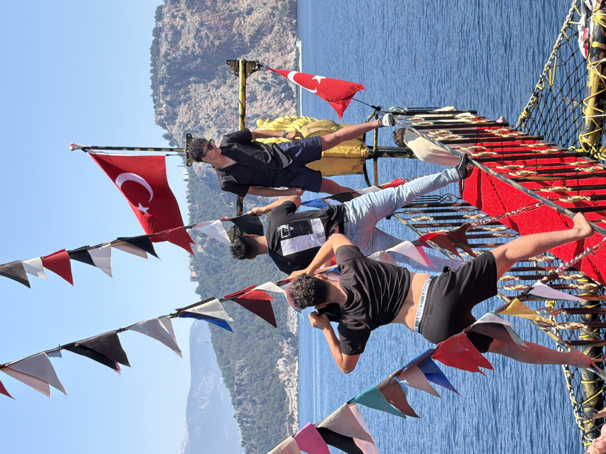 This media file captures a vibrant outdoor scene aboard a pirate-themed tour boat in Alanya, Turkey, under a clear blue sky on a bright, sunny day. Three young men are engaged in playful, acrobatic poses on the boat's rigging and decks.
The man on the left, wearing a black t-shirt, dark shorts, and sunglasses, is balancing on a wooden beam with his left leg extended horizontally, his right leg bent, and his arms held up, as if holding onto something out of frame. Behind him, a Turkish flag with a white crescent moon and star on a red background flies prominently from a mast.
The man in the middle, wearing a black t-shirt with a white design and light-colored jeans, stands on a lower platform, appearing to interact with or support the first man. The man on the right, dressed in a black t-shirt and black shorts, leans back with his legs extended upwards and his arms bent near his head, balancing on a red-carpeted structure resembling stairs or a slide. The elastic waistband of his shorts clearly shows the brand name "LACOSTE" and "SPORT".
The boat features decorative elements like a red carpet on the steps, gold accents on railings, and a safety net on the right. Colorful pennant flags (bunting) in shades of red, white, black, blue, and green are strung across the boat, adding to the festive atmosphere.
In the background, the serene blue water of the sea stretches out, meeting rugged, verdant mountains or cliffs under the expansive sky. The overall scene suggests a fun, adventurous activity or performance on a tourist excursion.