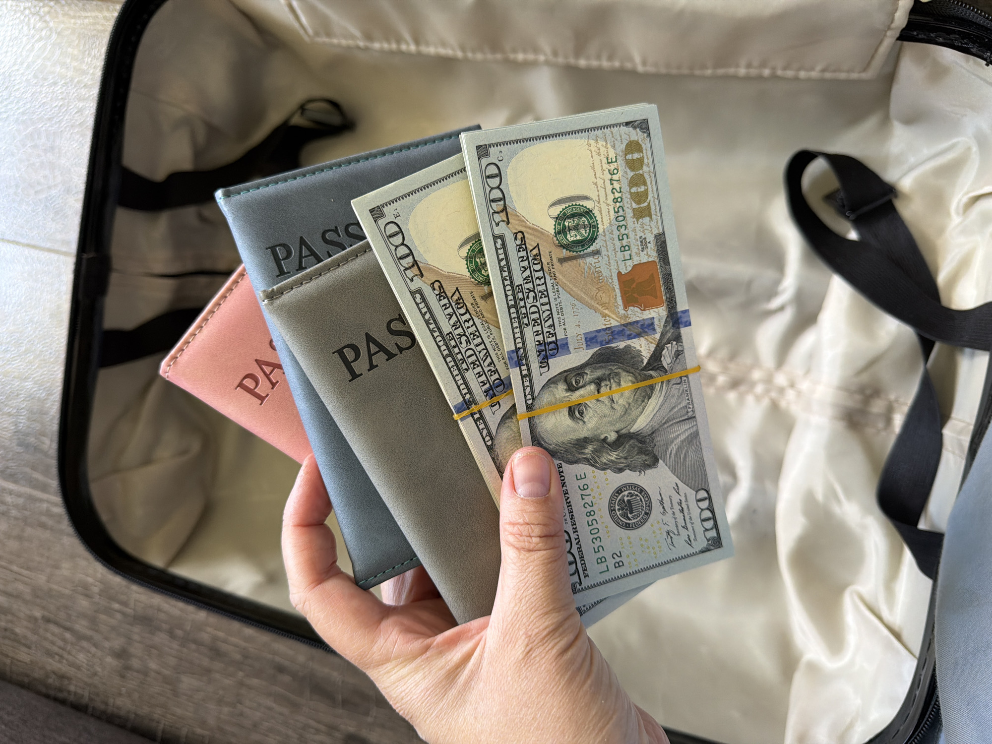A hand holds three passport covers and a stack of US hundred-dollar bills, bound together by a rubber band, over an open suitcase. The passport covers are colored pink, gray, and blue and are labeled "PASSPORT" in black. The suitcase has a beige interior lining and black straps. The background shows part of a wooden floor. No information about the city of Chaikovsky, Russia, can be gathered from the image.